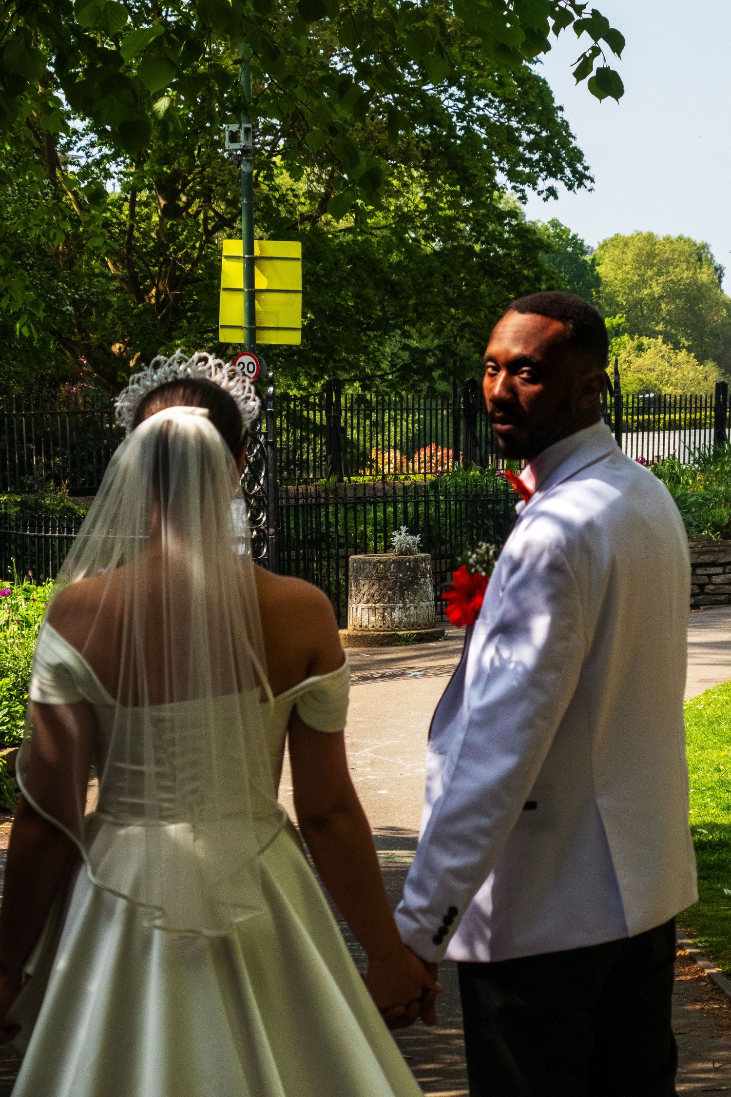 A couple in wedding attire walks along a garden path surrounded by lush green trees on a sunny day.