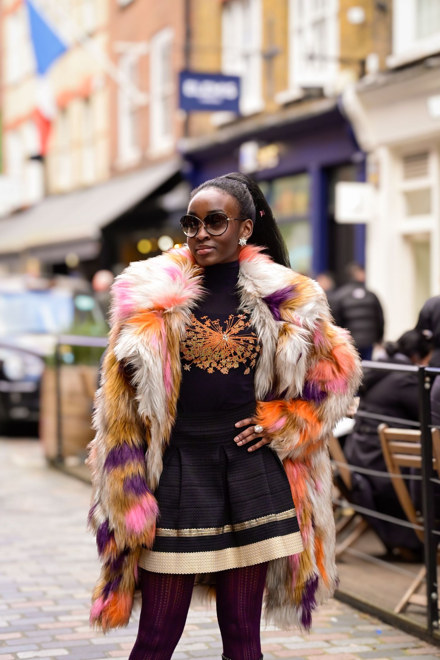 A colorful faux fur coat styled with a black dress and gold accessories against an urban backdrop.