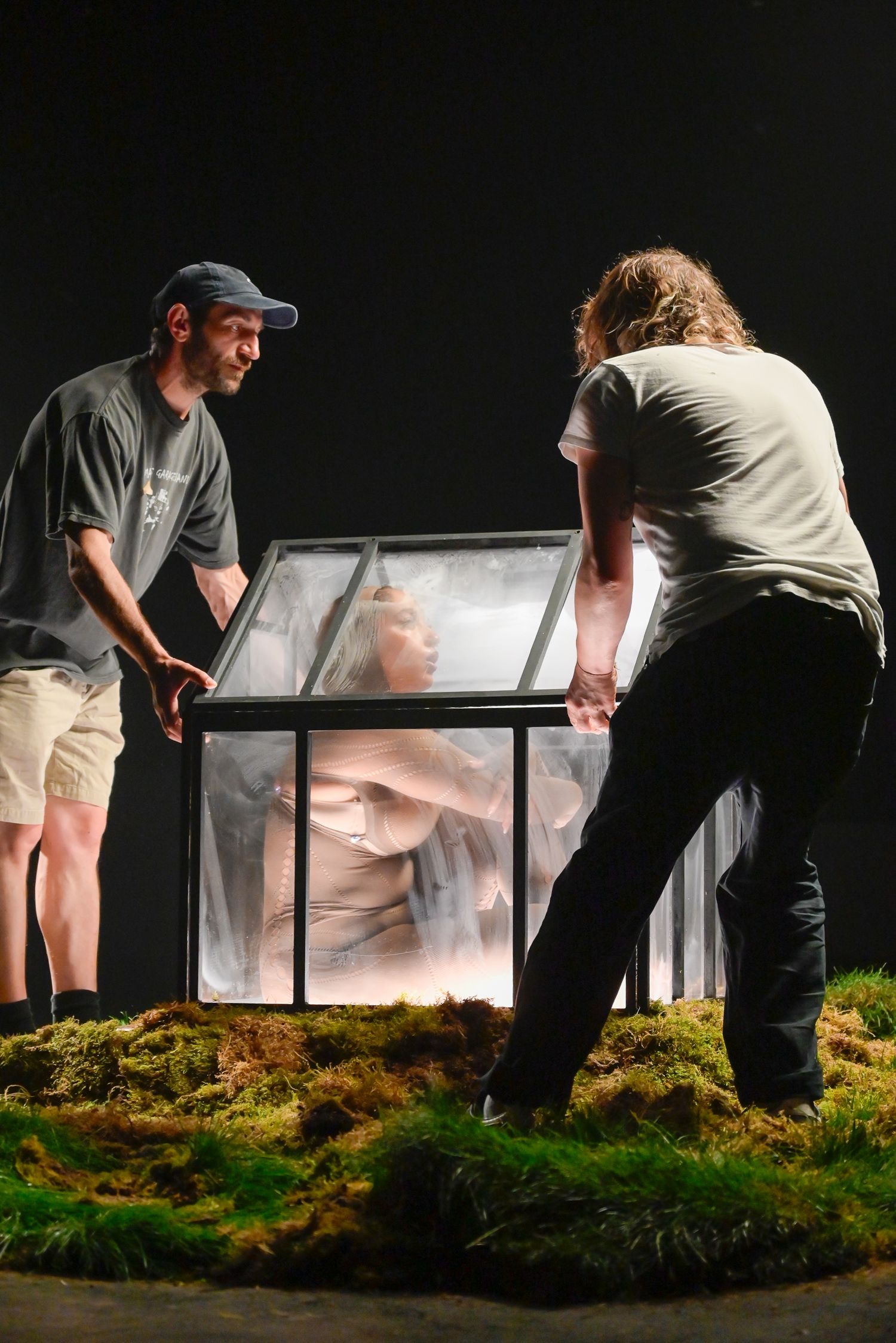 Behind-the-scenes setup of glass greenhouse art installation on textured moss base.