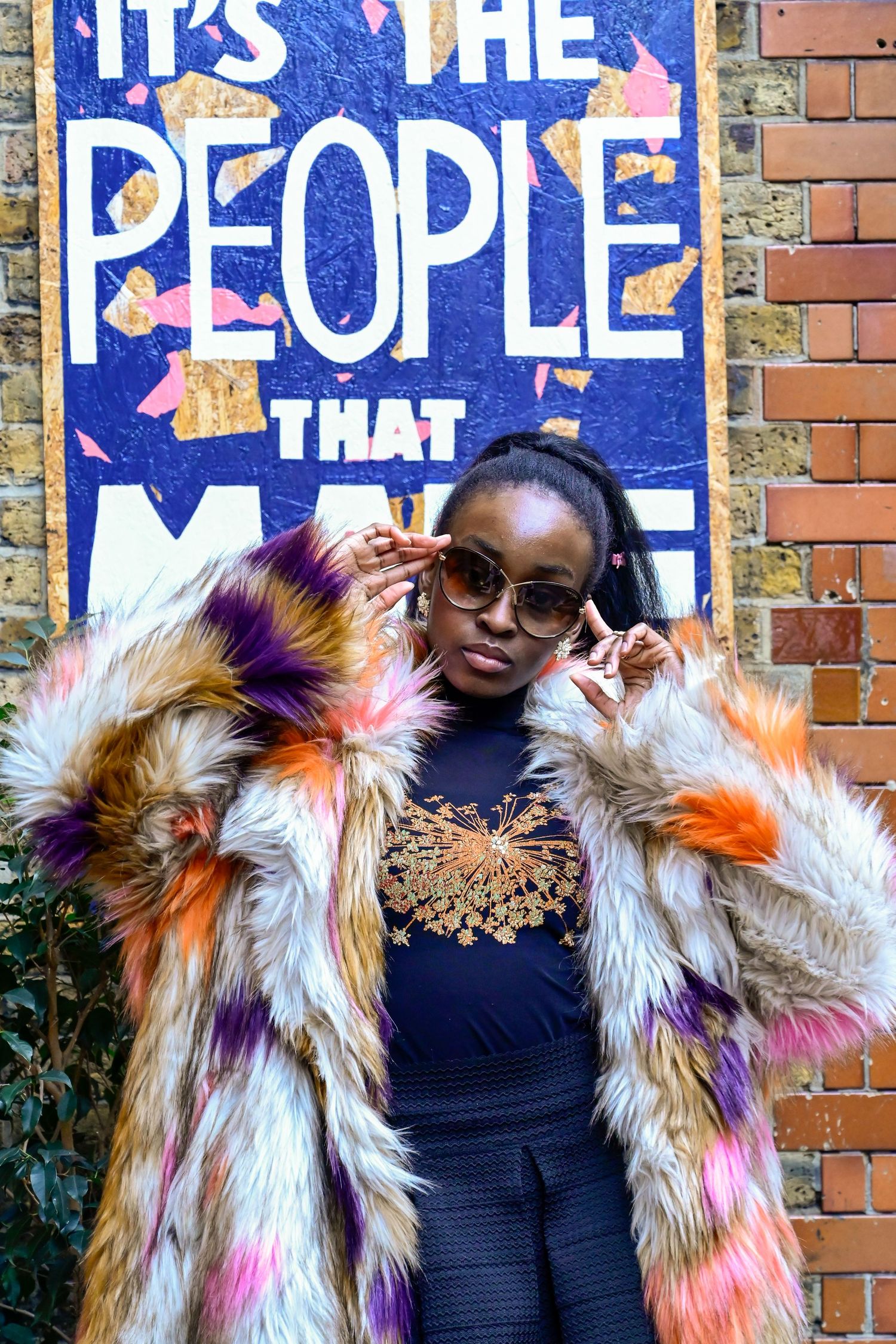 Statement multicolored fur coat shown against blue street art and brick wall background.