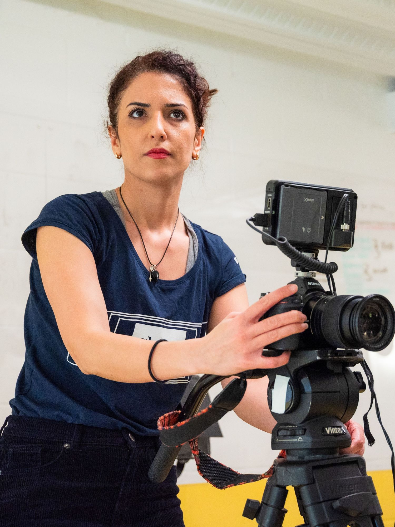 A person in a navy blue shirt adjusts a professional video camera mounted on a tripod against a light background.
