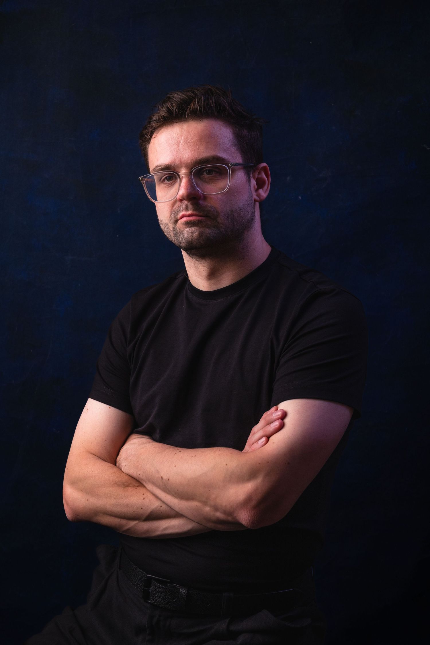 Professional portrait of someone in a black t-shirt with crossed arms against a dark blue backdrop.