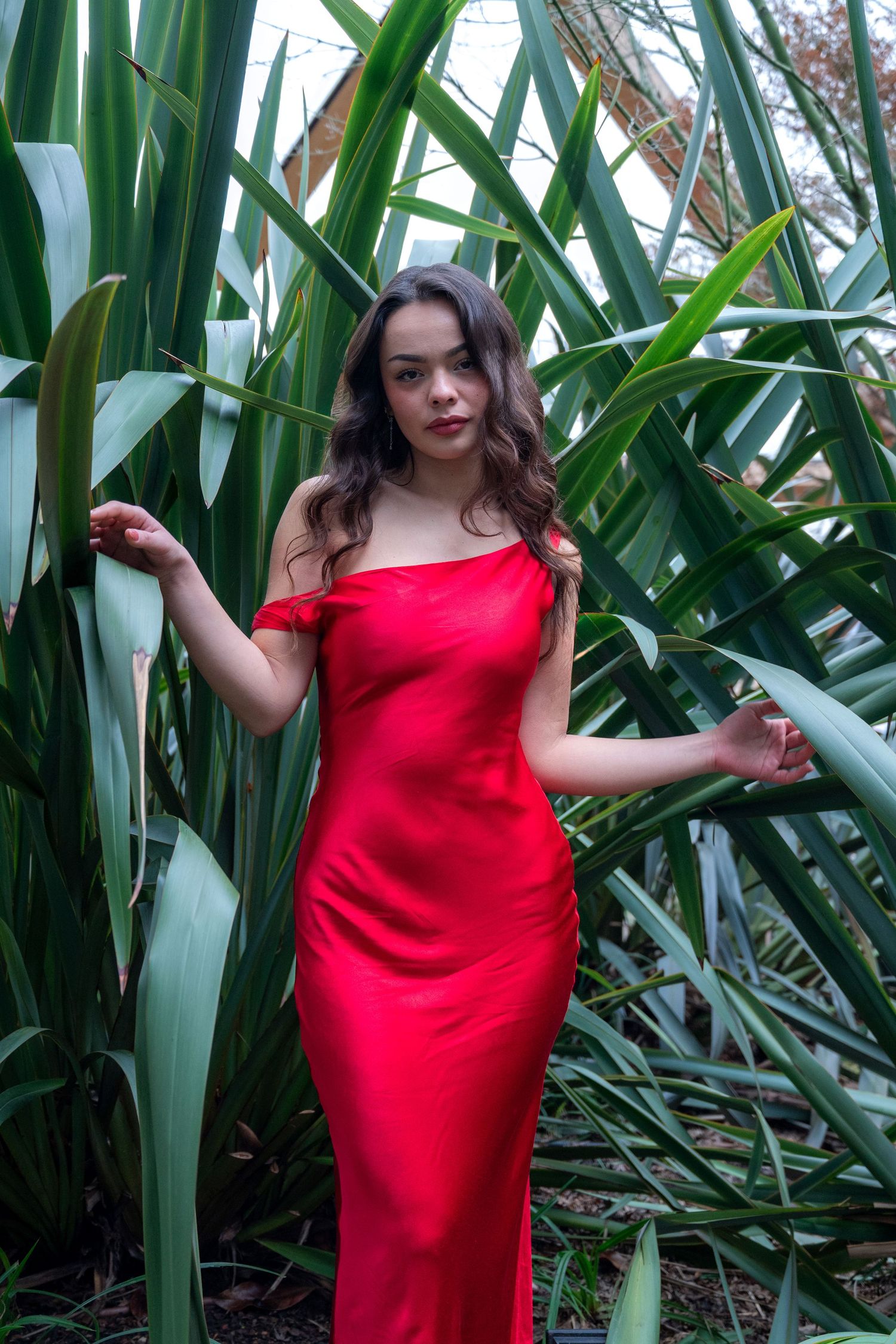 Woman in elegant red off-shoulder satin gown posing among tall green tropical plants outdoors.