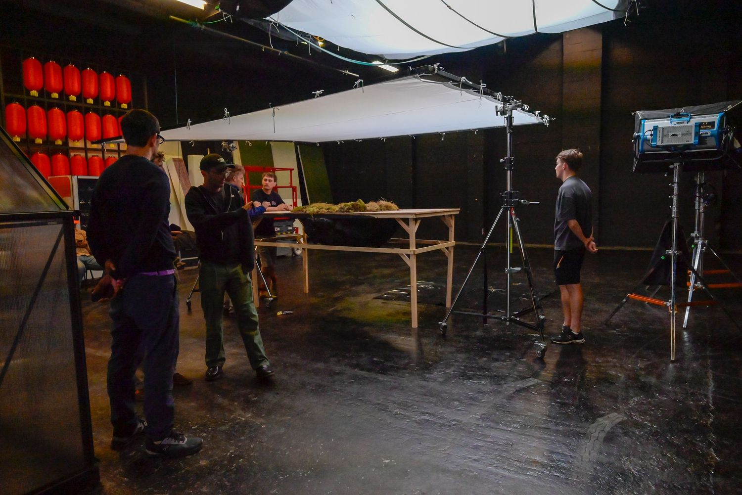 Production crew setting up equipment and lighting around a table in dark studio space.