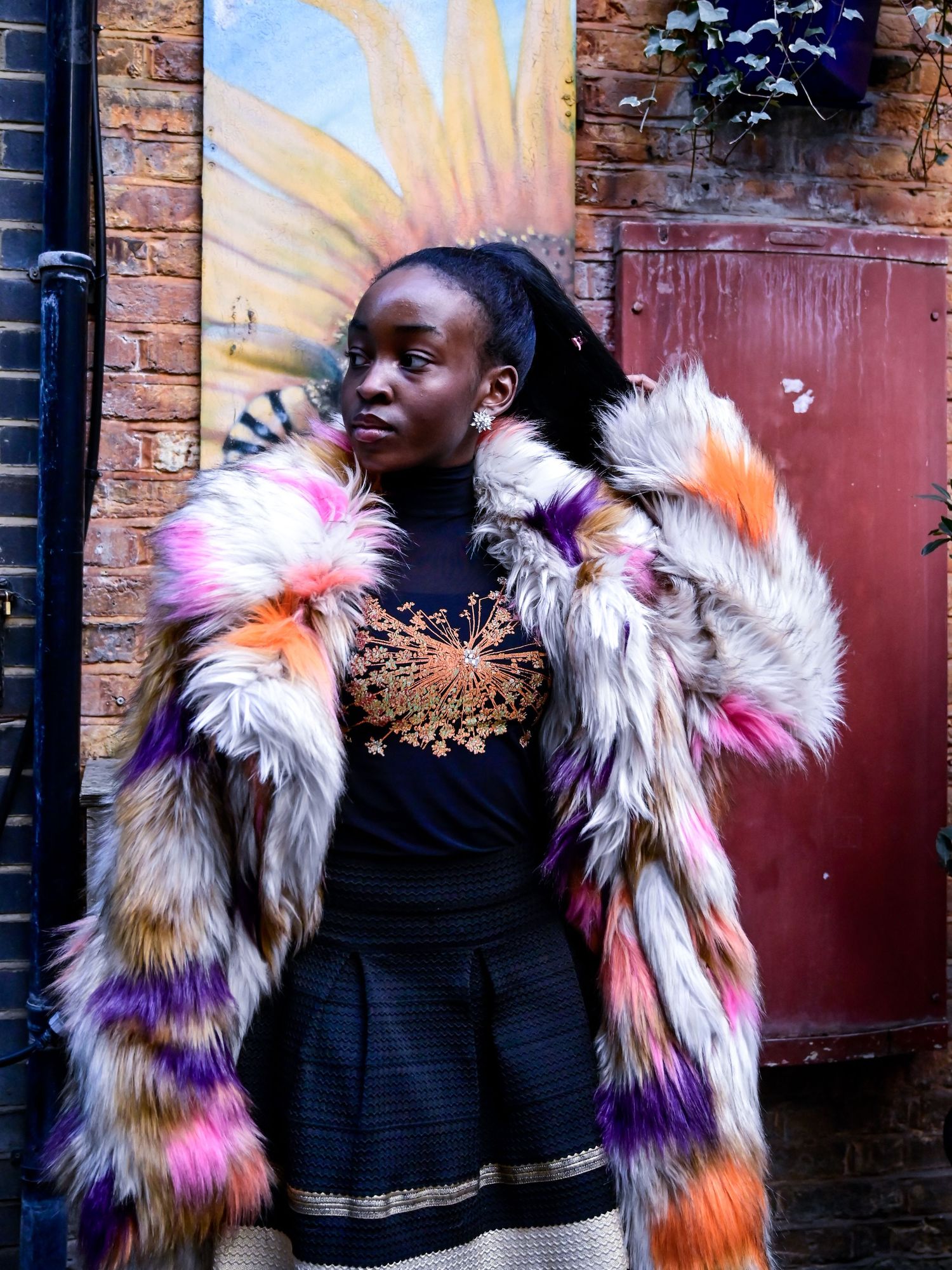 Multicolored furry jacket with pink, orange and purple tones paired with a black outfit against brick wall.