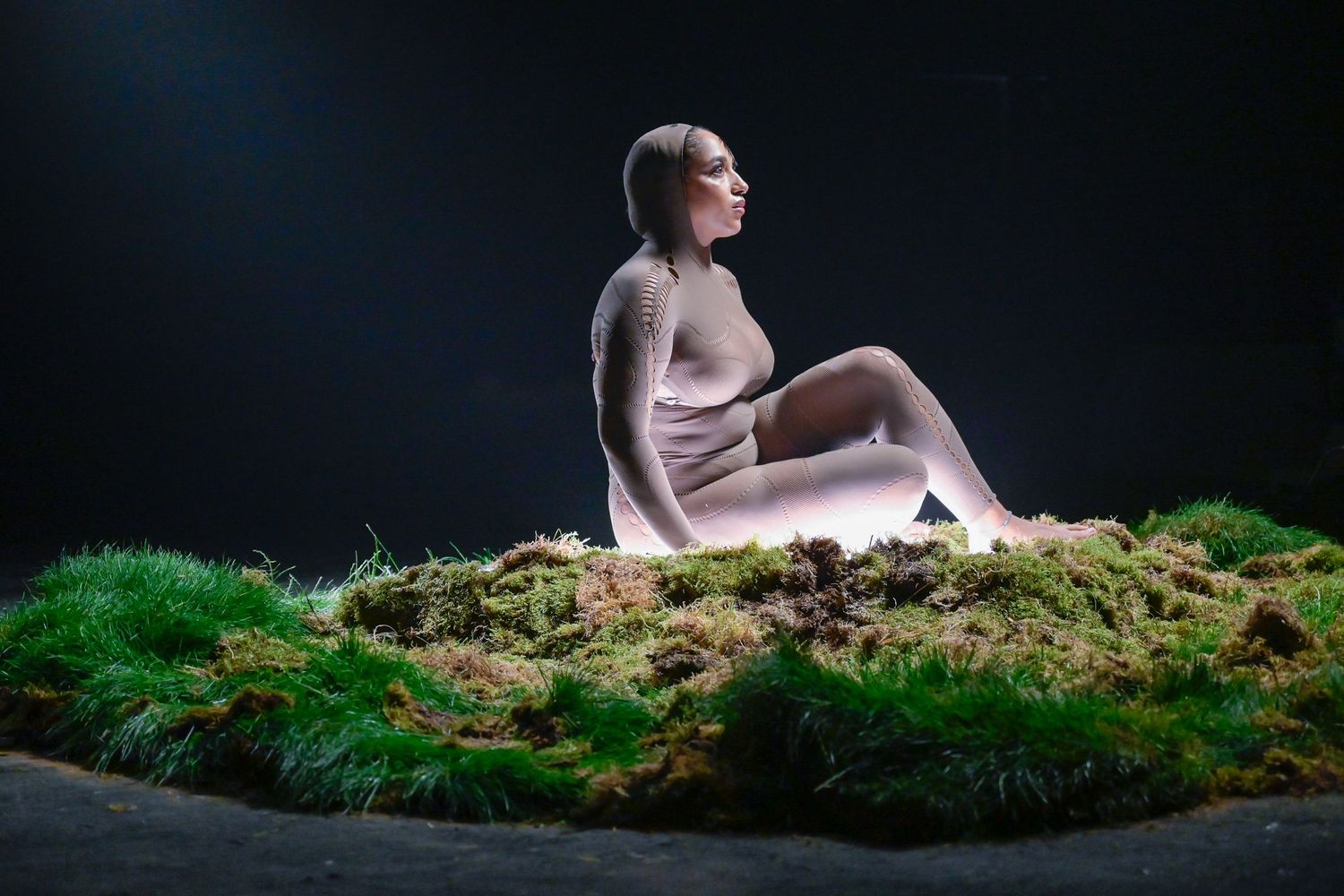 Artistic photo of a figure seated on moss-covered ground against a dark background with dramatic lighting.
