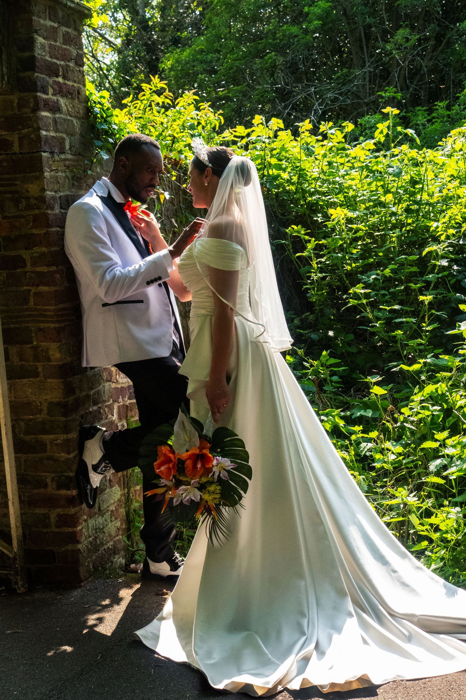 Newlyweds in elegant wedding attire pose against brick wall surrounded by lush garden.