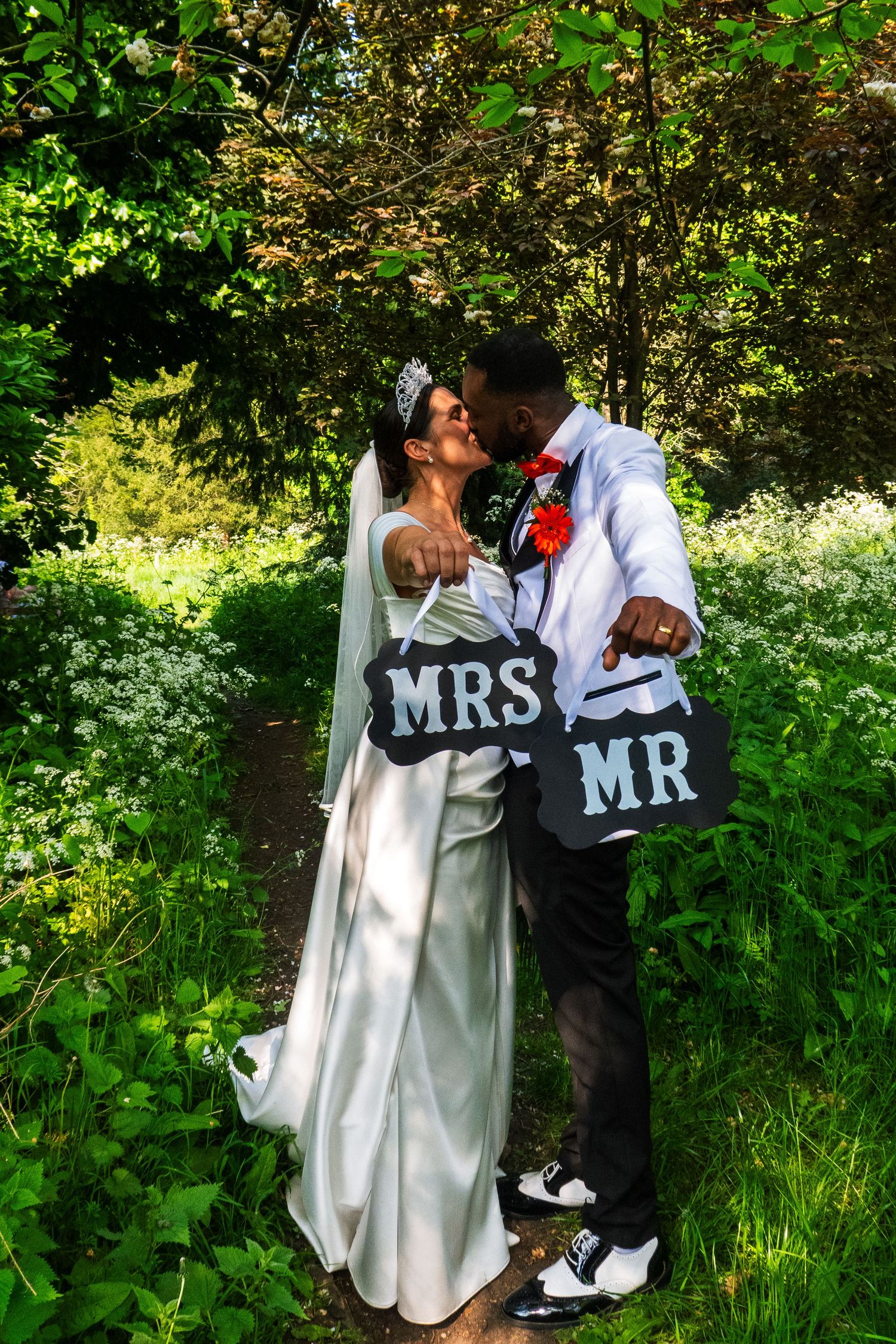 Newlyweds share an intimate moment while holding Mr and Mrs signs in a green garden setting.