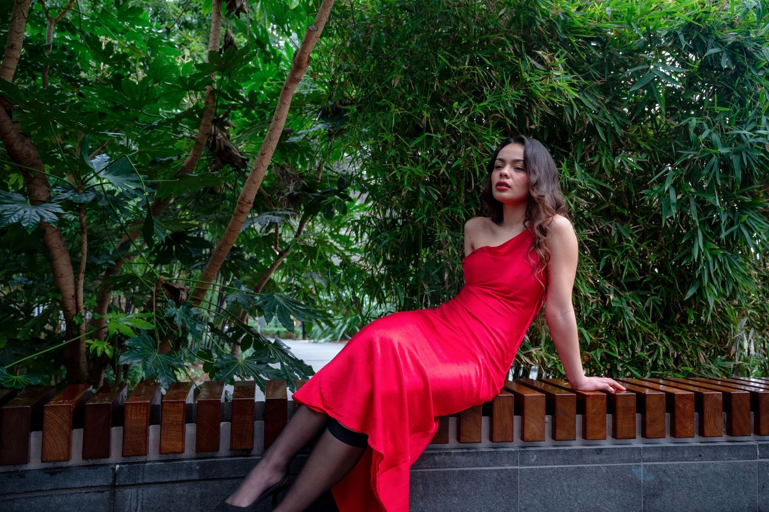 Woman in red dress sitting on fence surrounded by lush green trees.