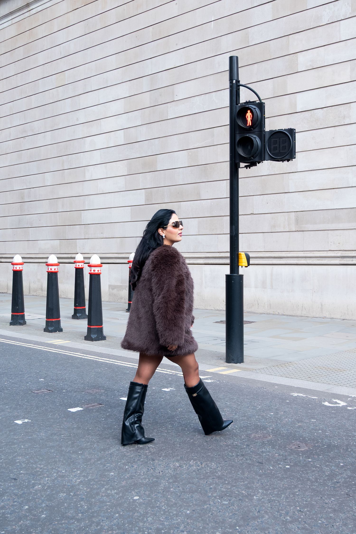 A person in a brown fur coat and black boots walks past a traffic light and bollards on a gray urban street.