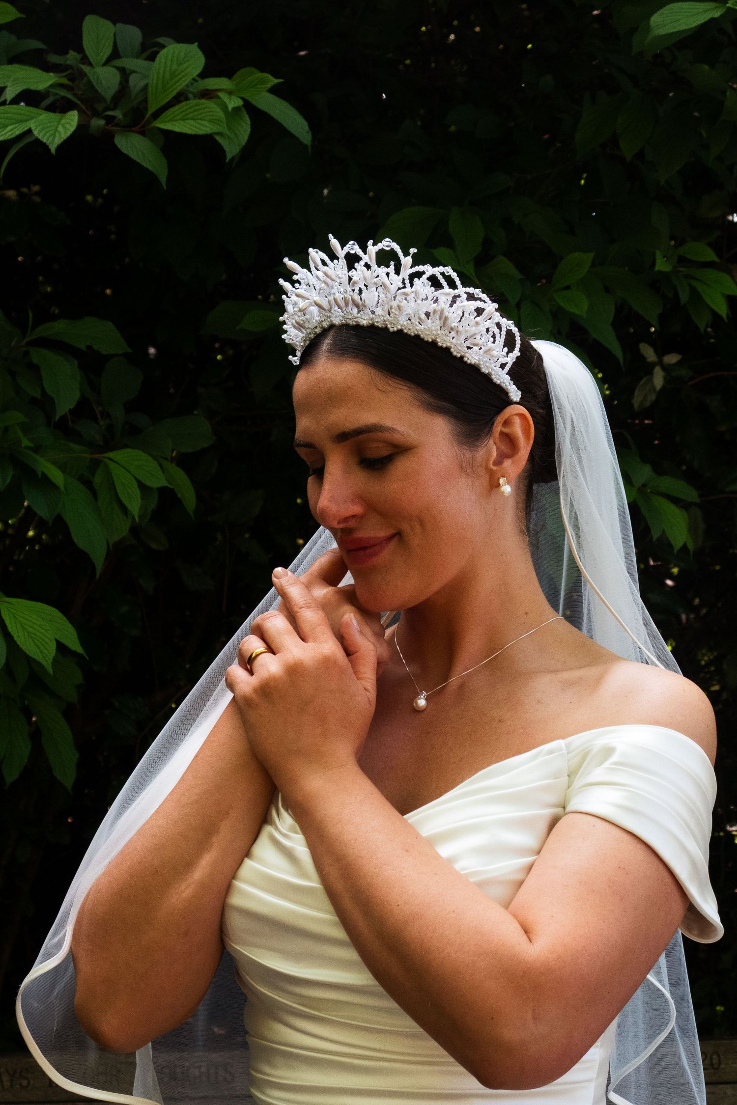 A bride wearing an off-shoulder dress and crystal tiara poses in front of green foliage.