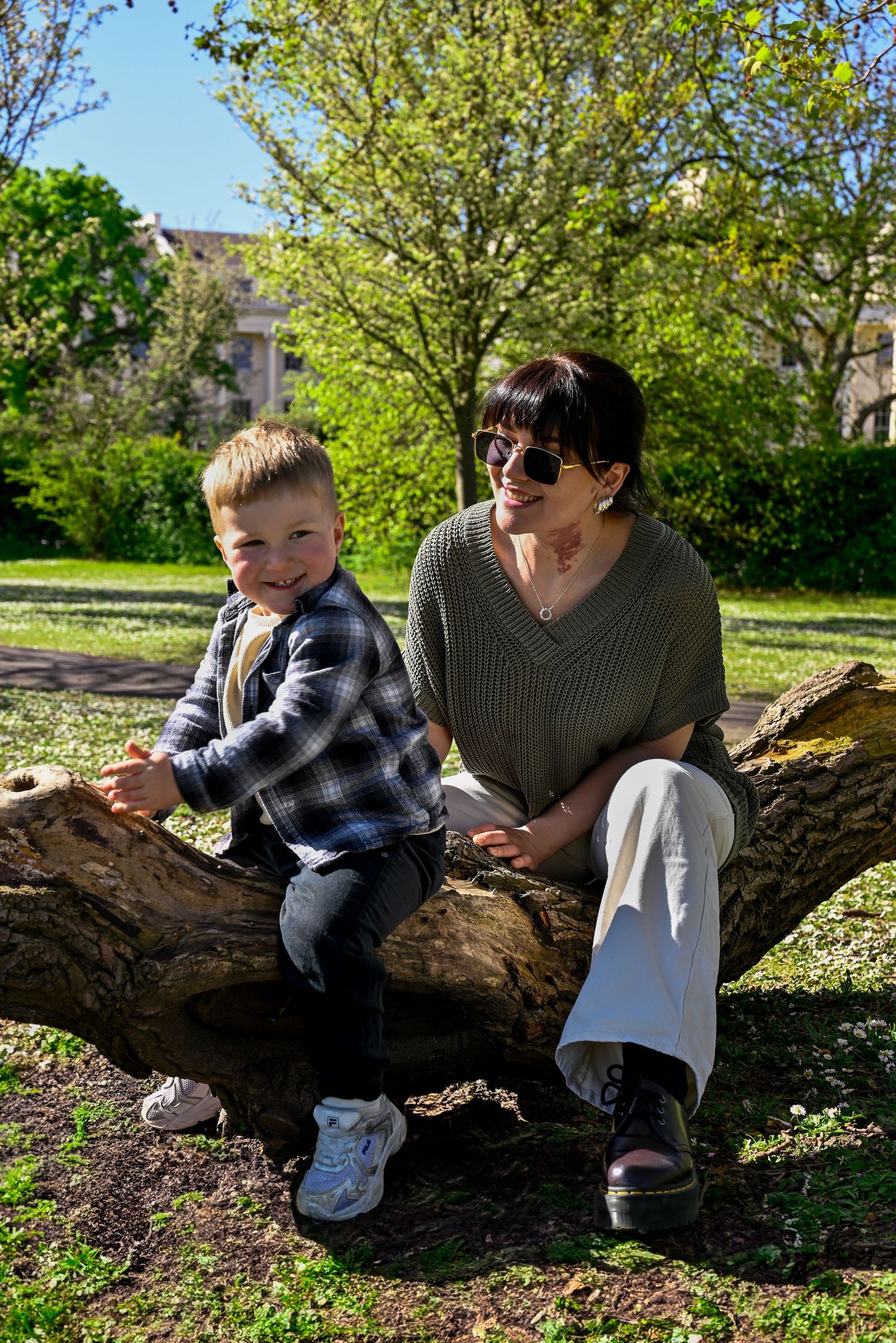 A woman and young boy sit together on a large tree log in a sunny park surrounded by green trees.