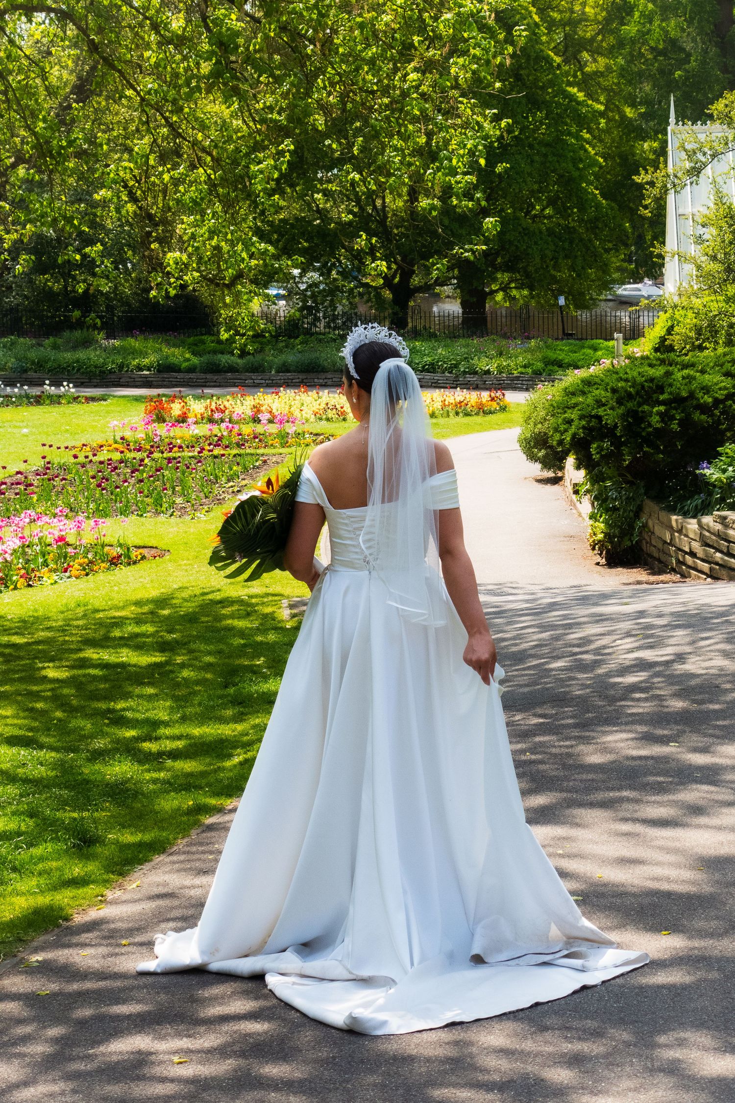 Two white wedding dresses flow elegantly along a garden path surrounded by lush greenery and colorful flowers.