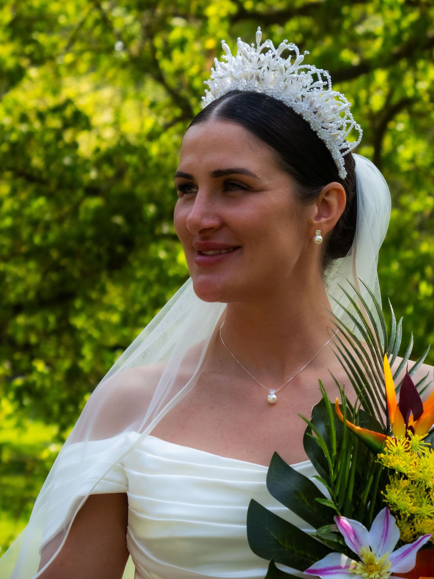A bride wearing a crystal tiara and veil holds a tropical bouquet against a green garden backdrop.