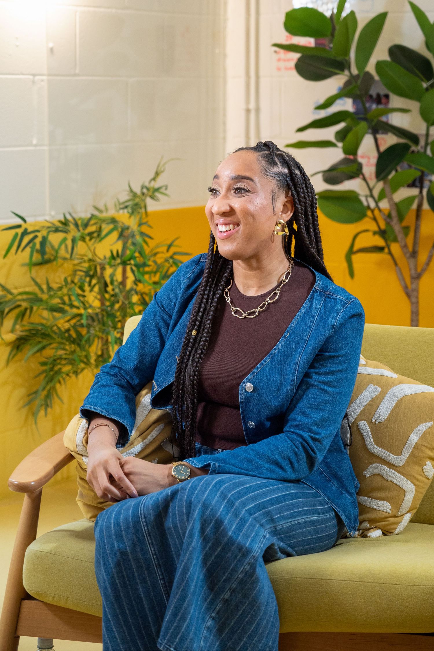 A person wearing a denim jacket sits casually on a green couch with indoor plants in the background.