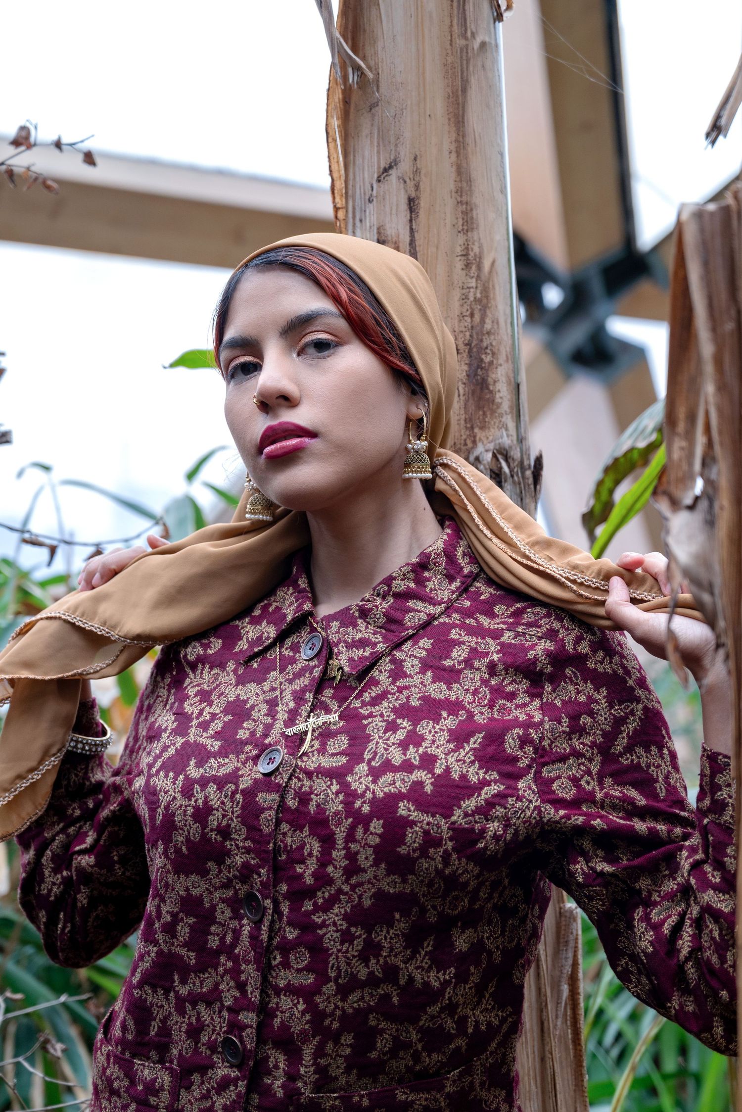 A young woman in a burgundy floral dress and tan headscarf poses gracefully among wooden posts and green foliage.