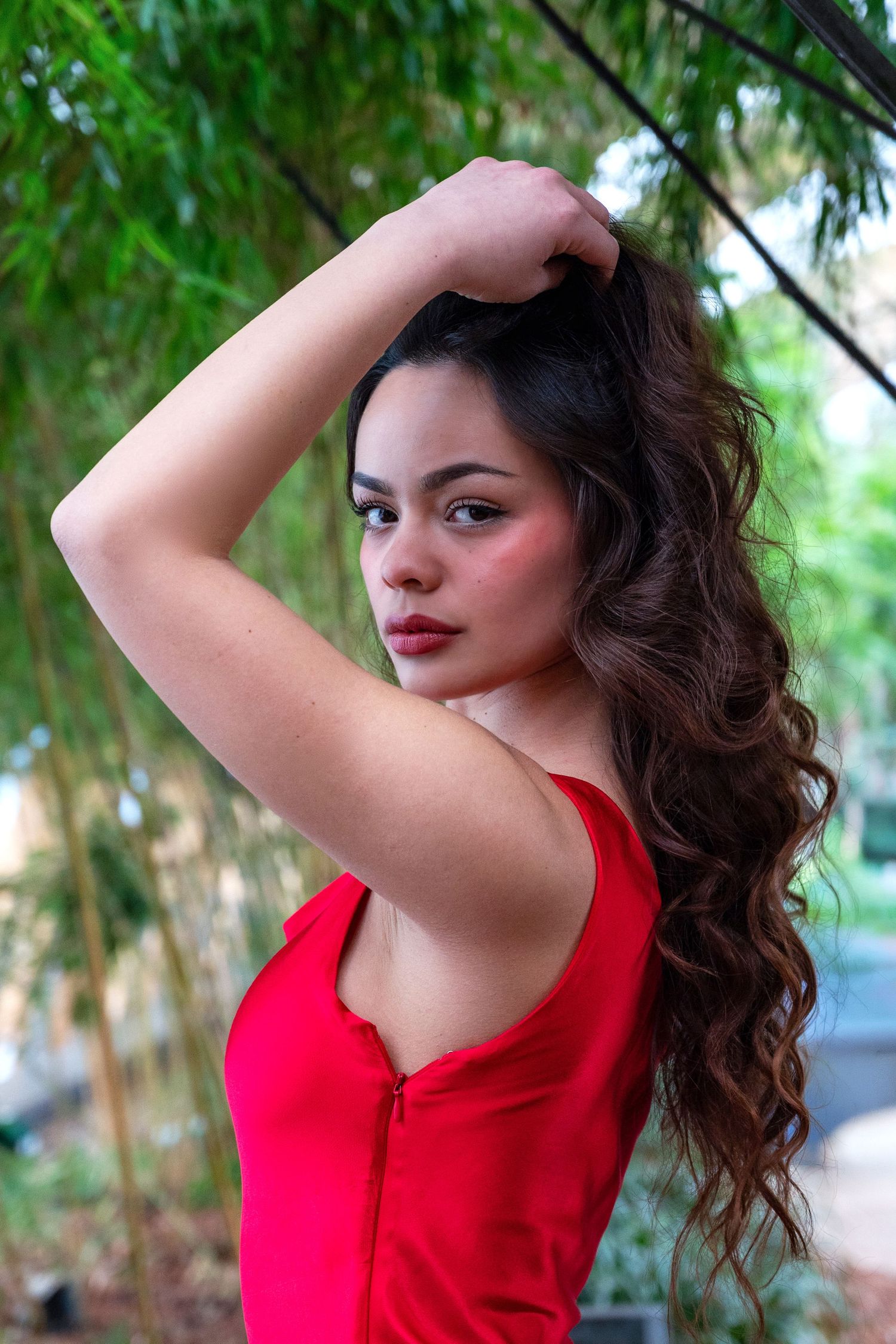 Young woman in red dress poses outdoors with hand in long curly brown hair, green bamboo background.