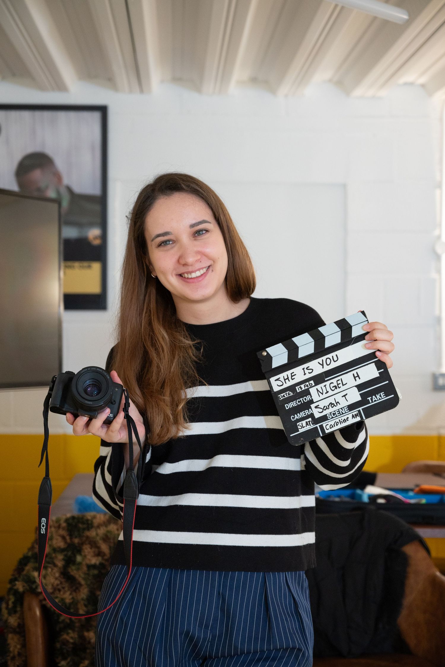 Smiling woman holding a film clapperboard and camera, wearing a striped sweater, standing in a creative studio space.