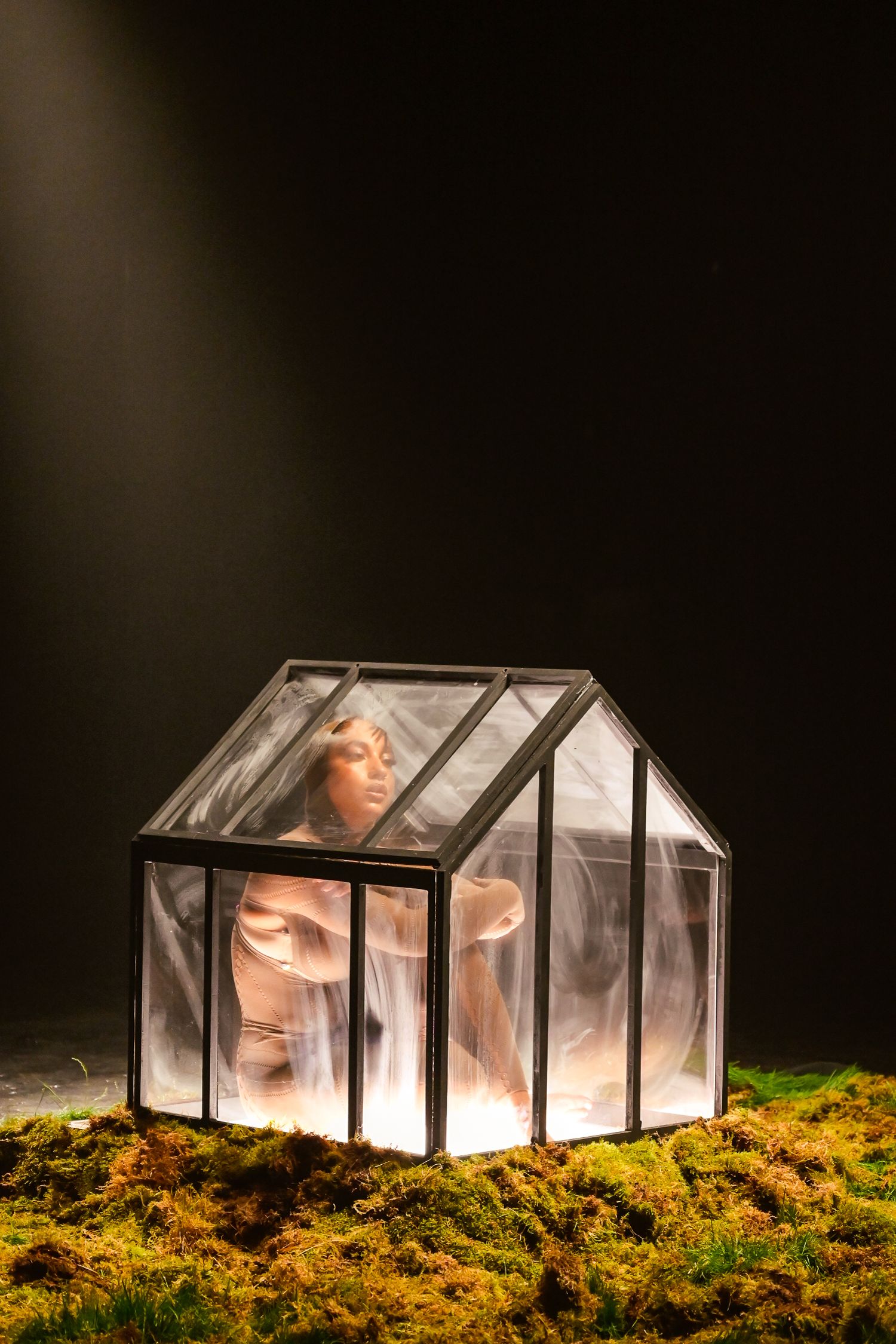Illuminated glass greenhouse structure on mossy ground creating ethereal nighttime scene.