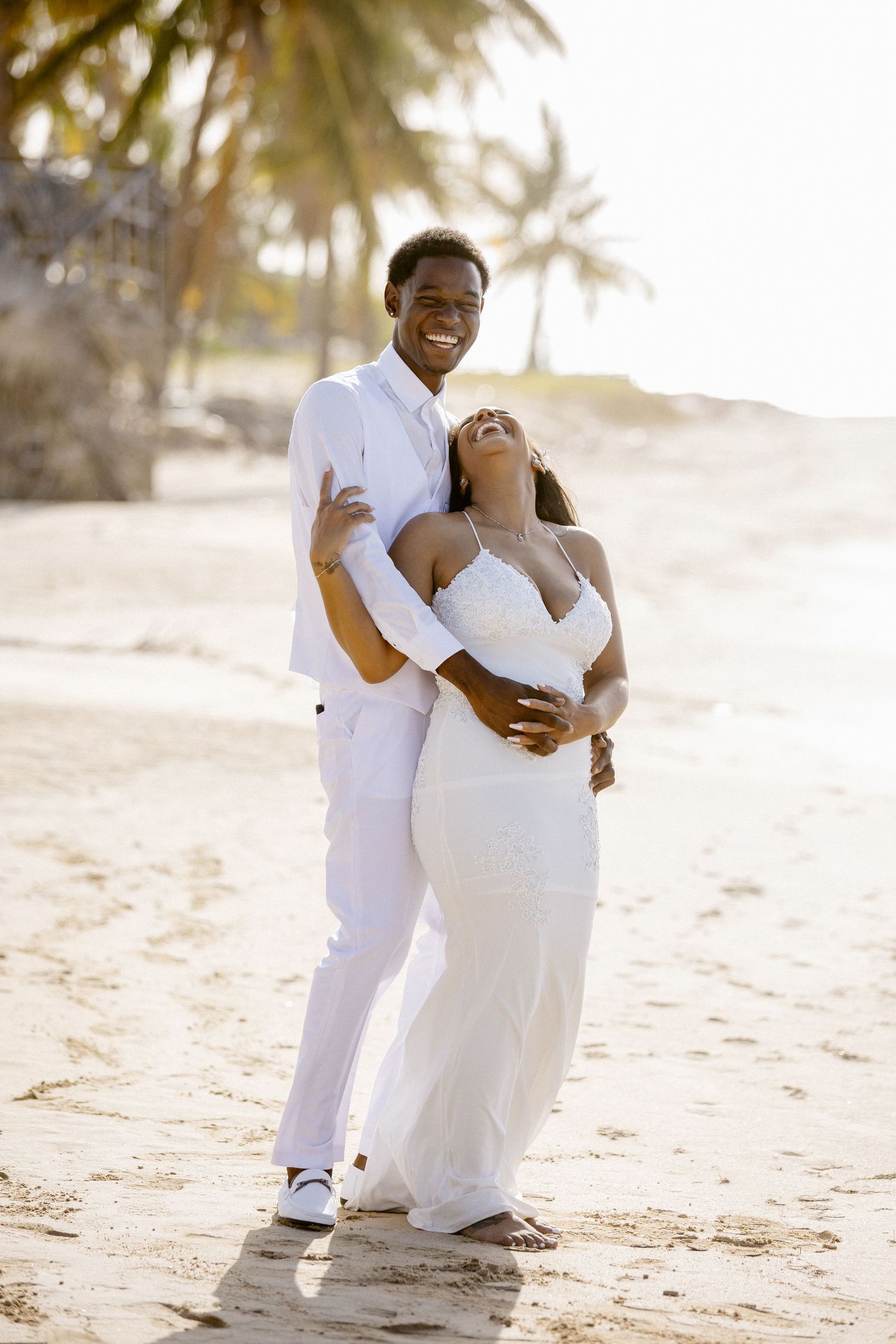 A happy couple embraces on a sunny beach, both dressed in white, surrounded by palm trees.