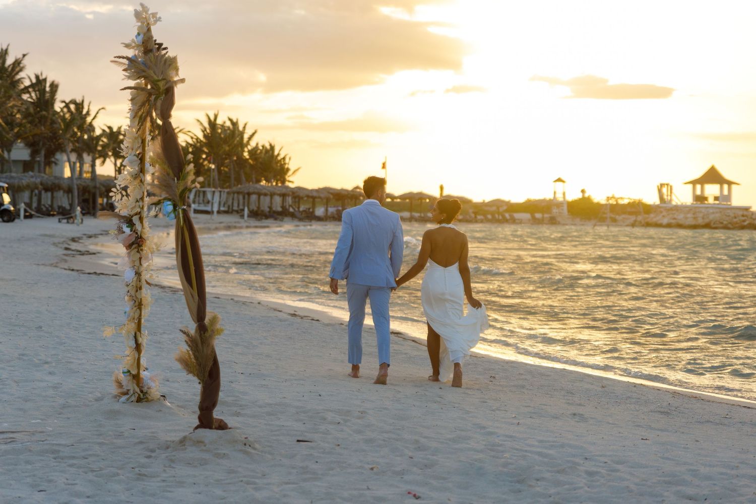 Couple walks hand-in-hand on tropical beach at sunset near floral wedding arch.