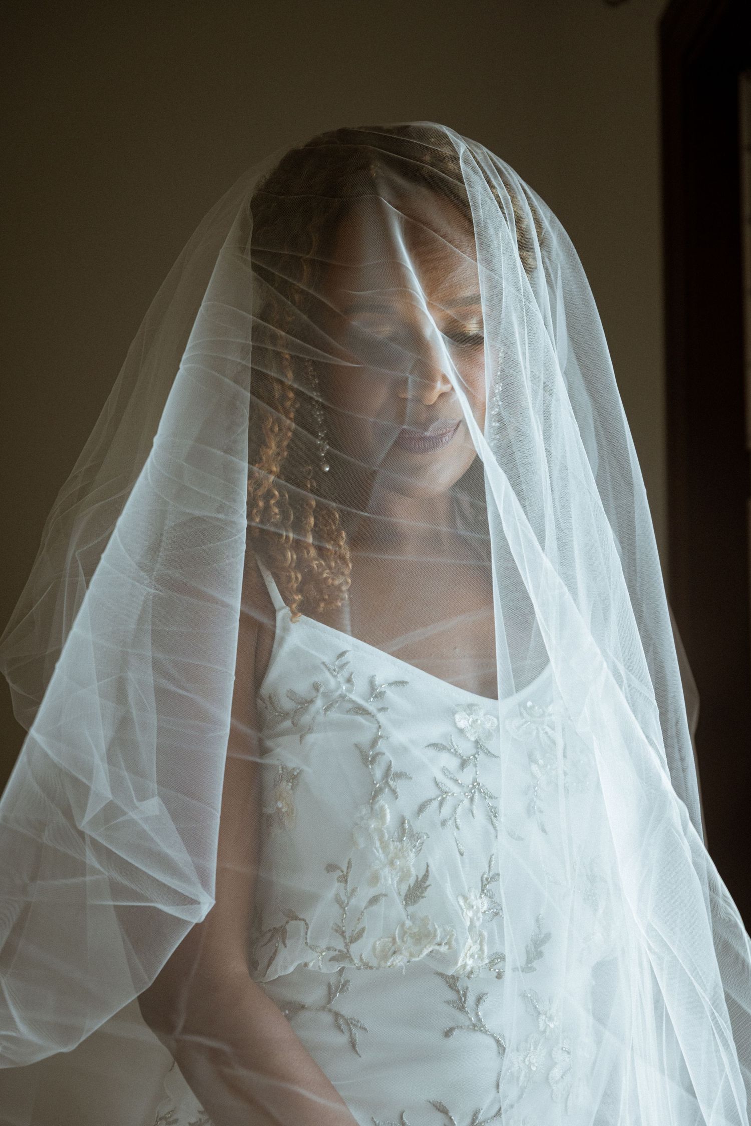 A bride with eyes closed smiles softly beneath a delicate white wedding veil in soft natural light.