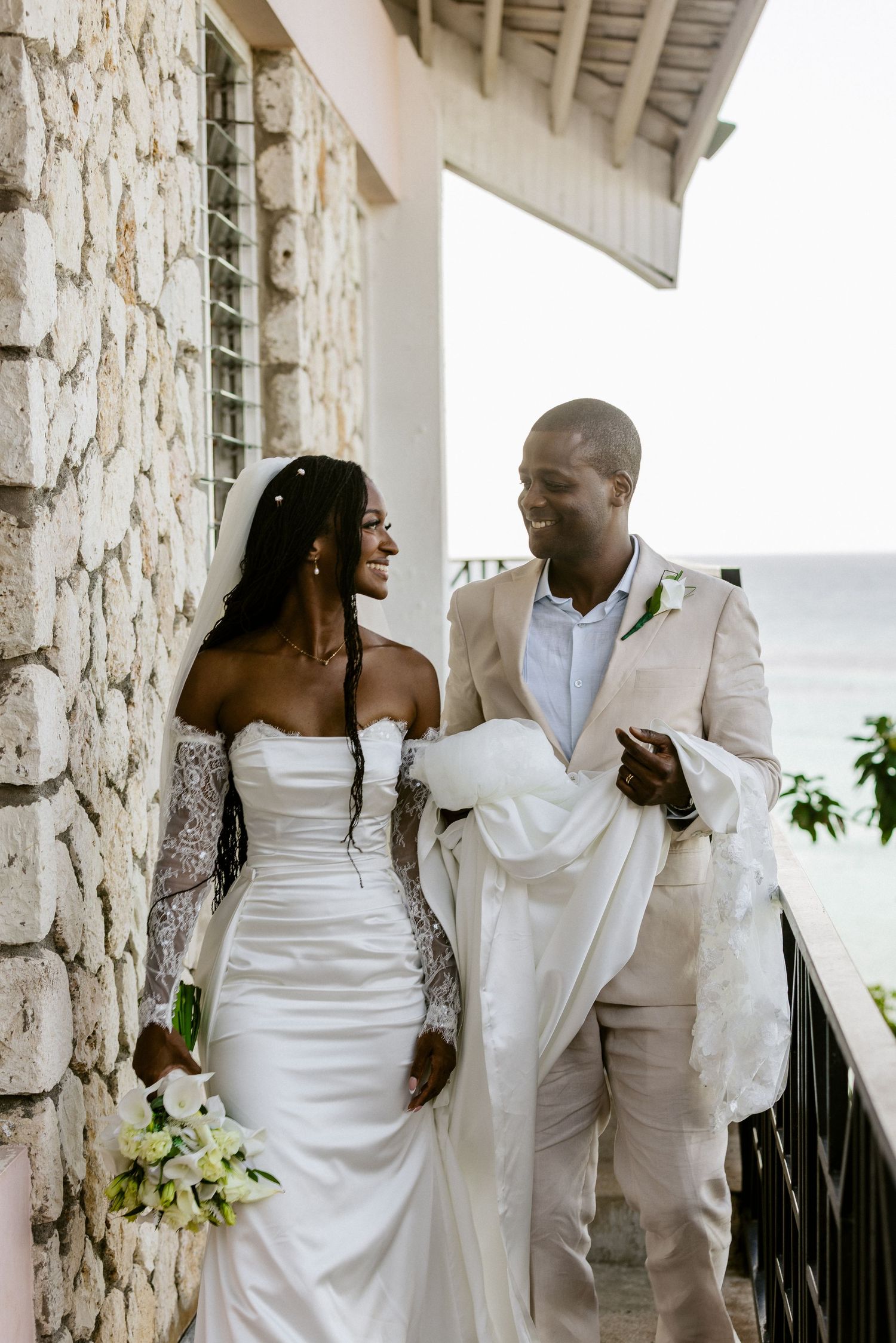 A smiling bride and groom walk together on a balcony by the ocean, bride in strapless gown with veil.