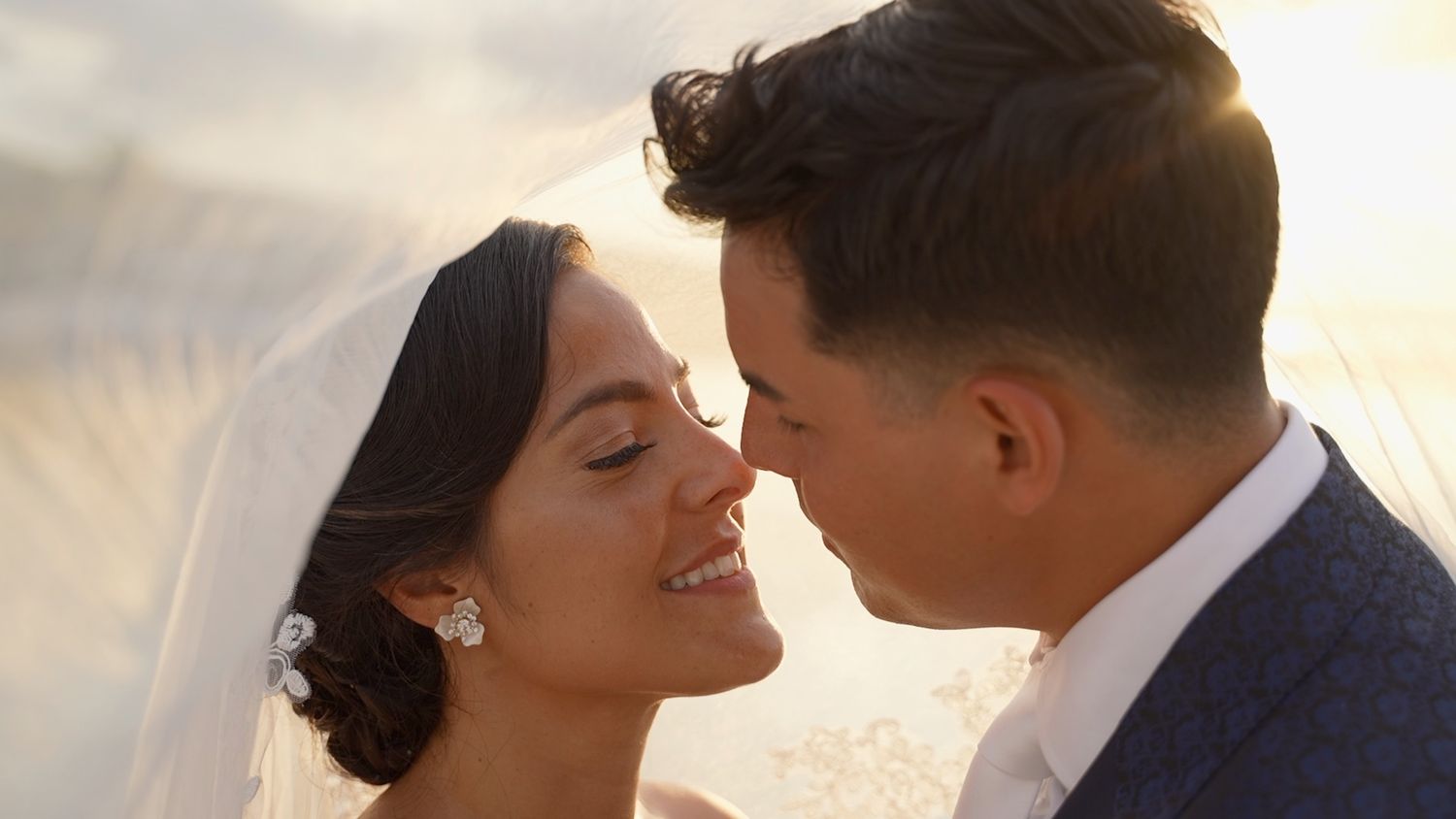 Bride and groom nearly kissing under veil with golden backlight at outdoor wedding.
