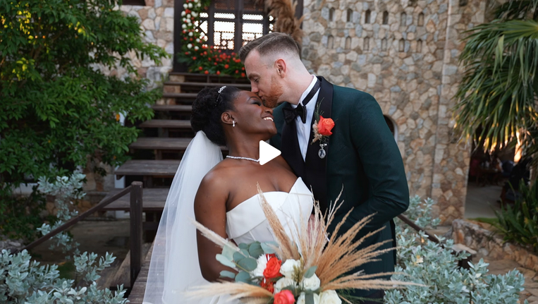 Bride and groom kissing in front of rustic stone building with colorful wedding bouquet.