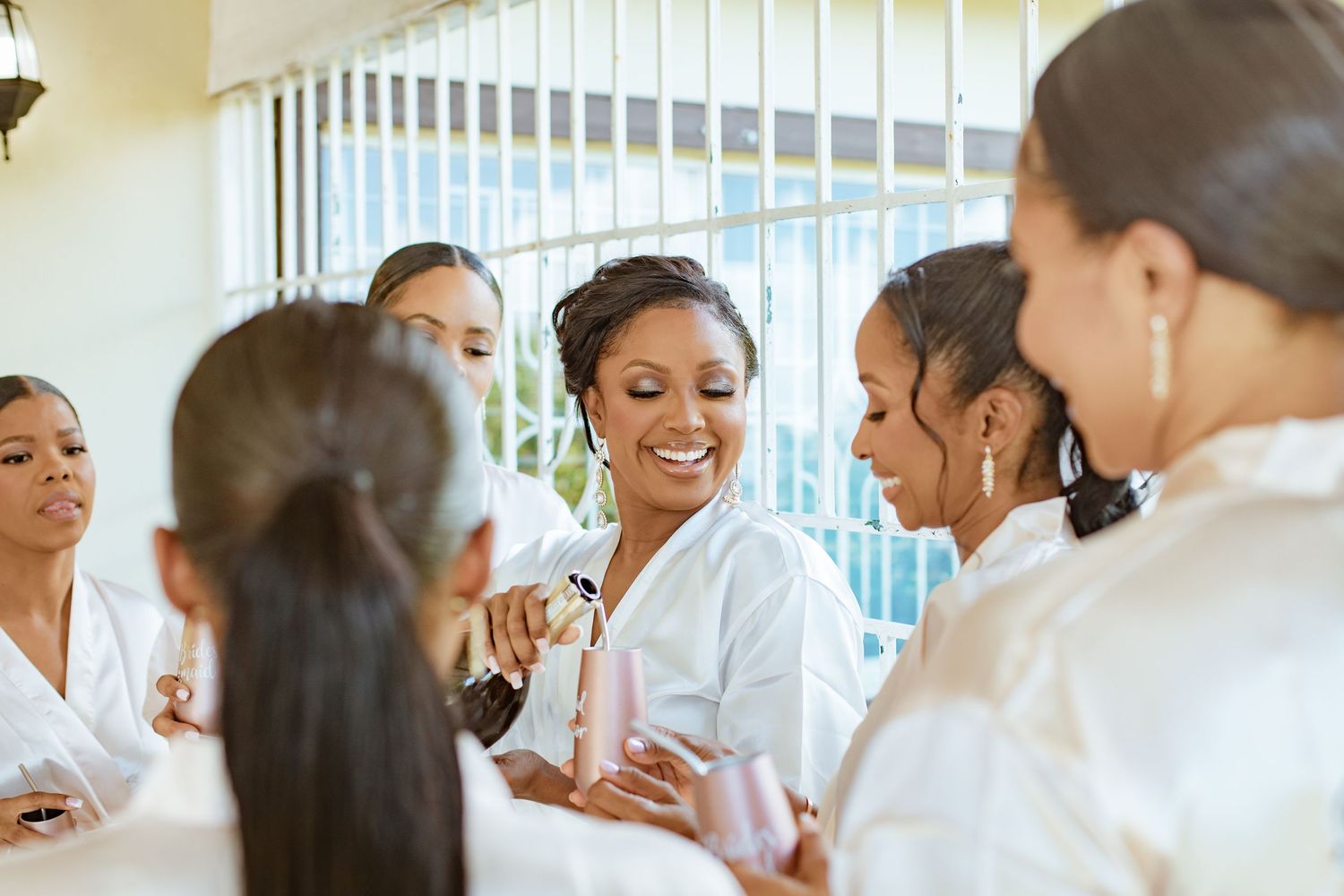 Bride smiling joyfully with bridesmaids in white robes during wedding morning preparations.