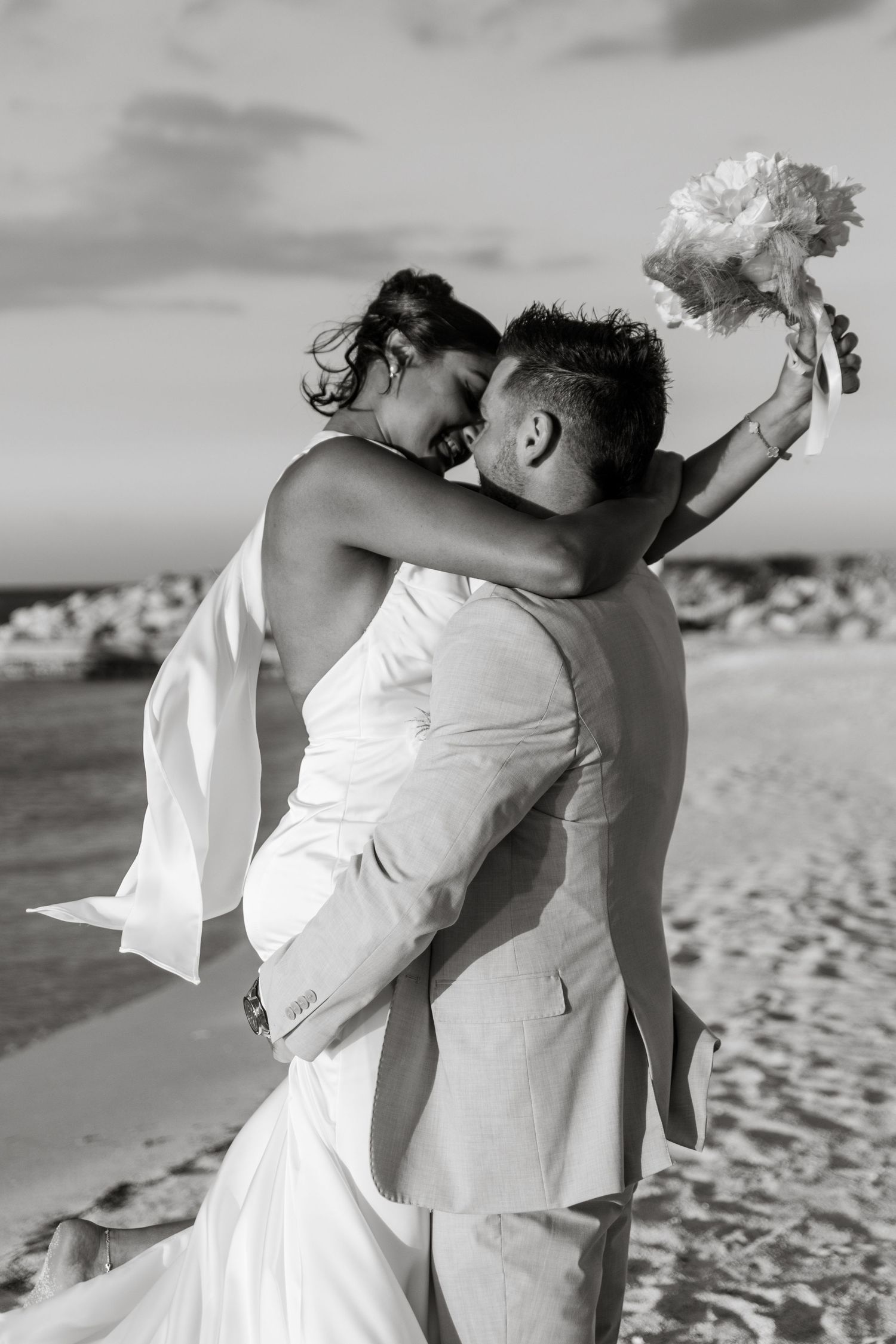 Black and white photo of groom lifting bride on beach holding bouquet.