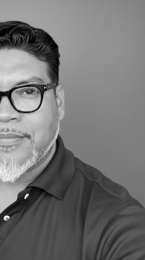 A black and white headshot of a man with thick-framed glasses and a salt-and-pepper goatee in a polo shirt.
