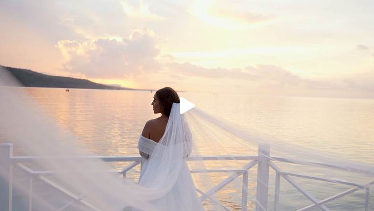 A bride in a flowing white gown and veil stands on a dock at sunset overlooking a calm tropical sea.