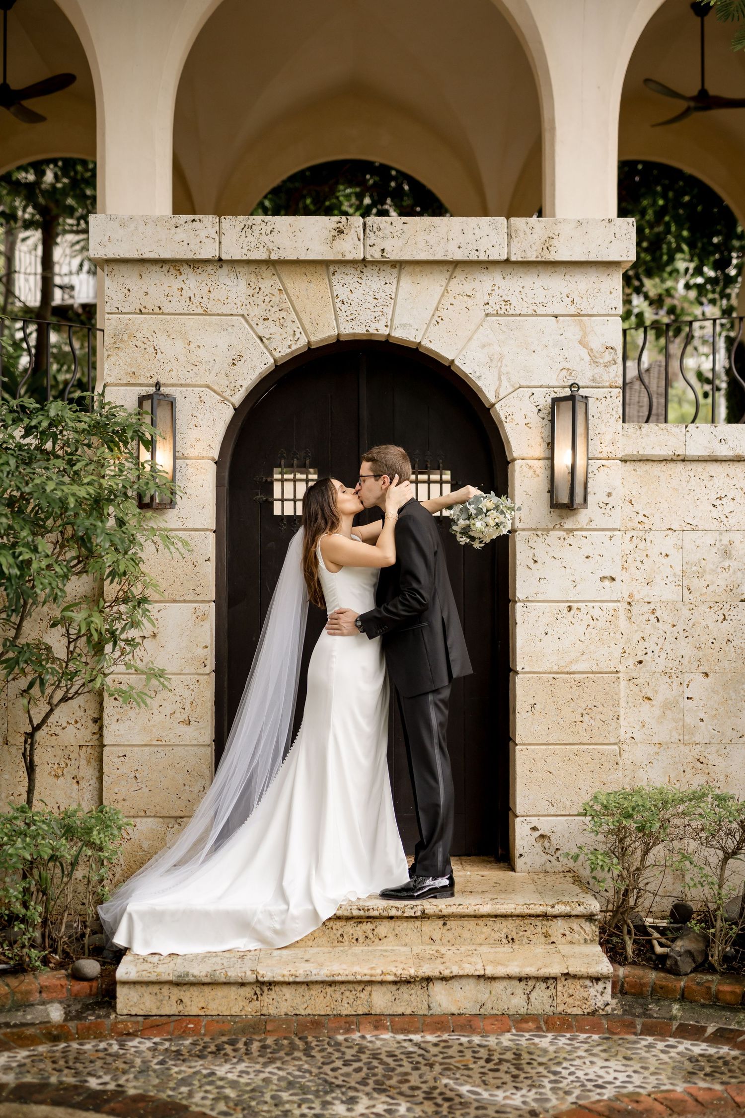 Bride and groom share a kiss on stone steps before an elegant arched wooden door at their wedding venue at Las Catalinas, Guanacaste.