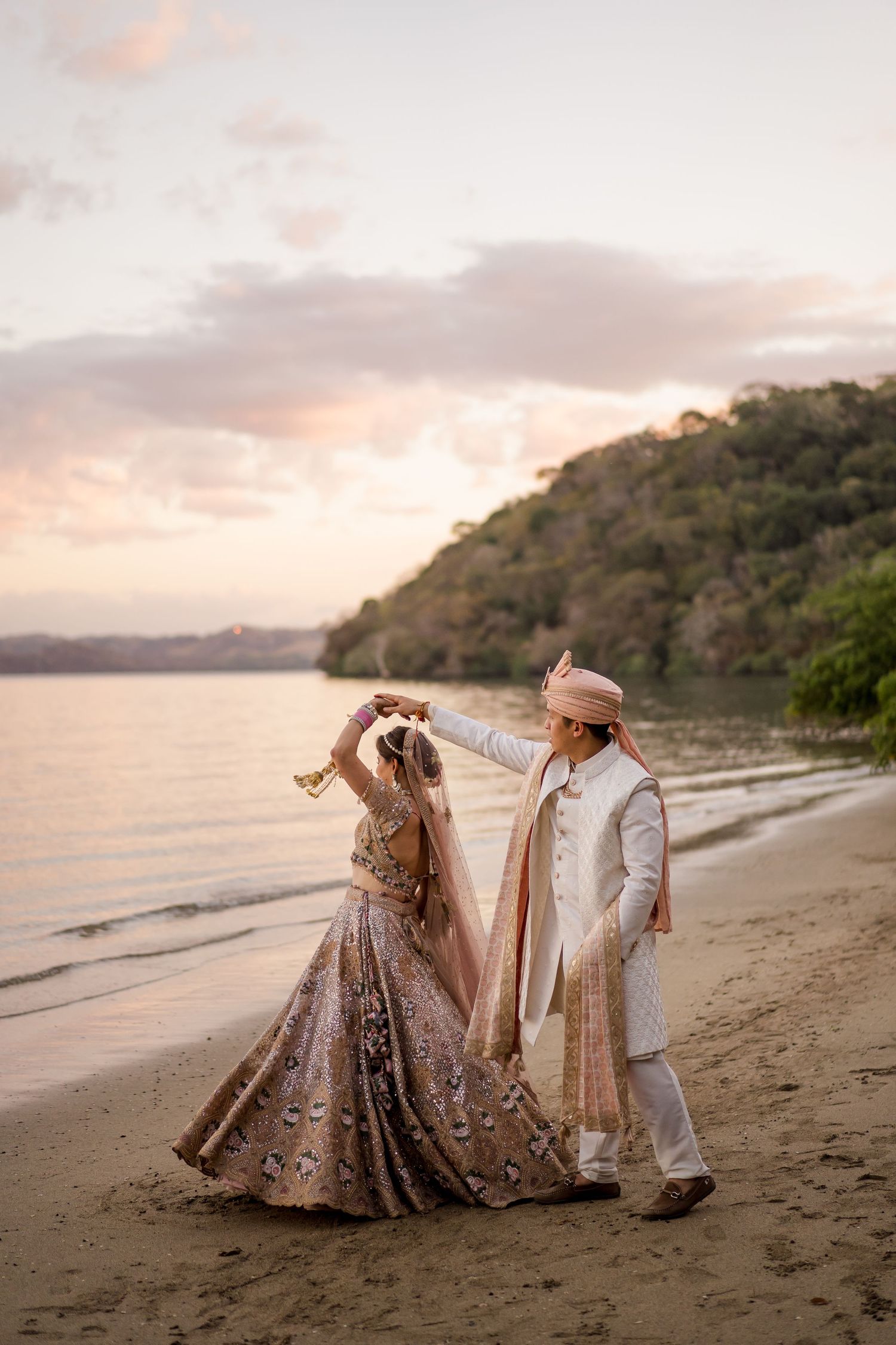 Indian bride and groom in traditional attire dancing on a beach at sunset with lush hills in the background in Guanacaste, Costa Rica.