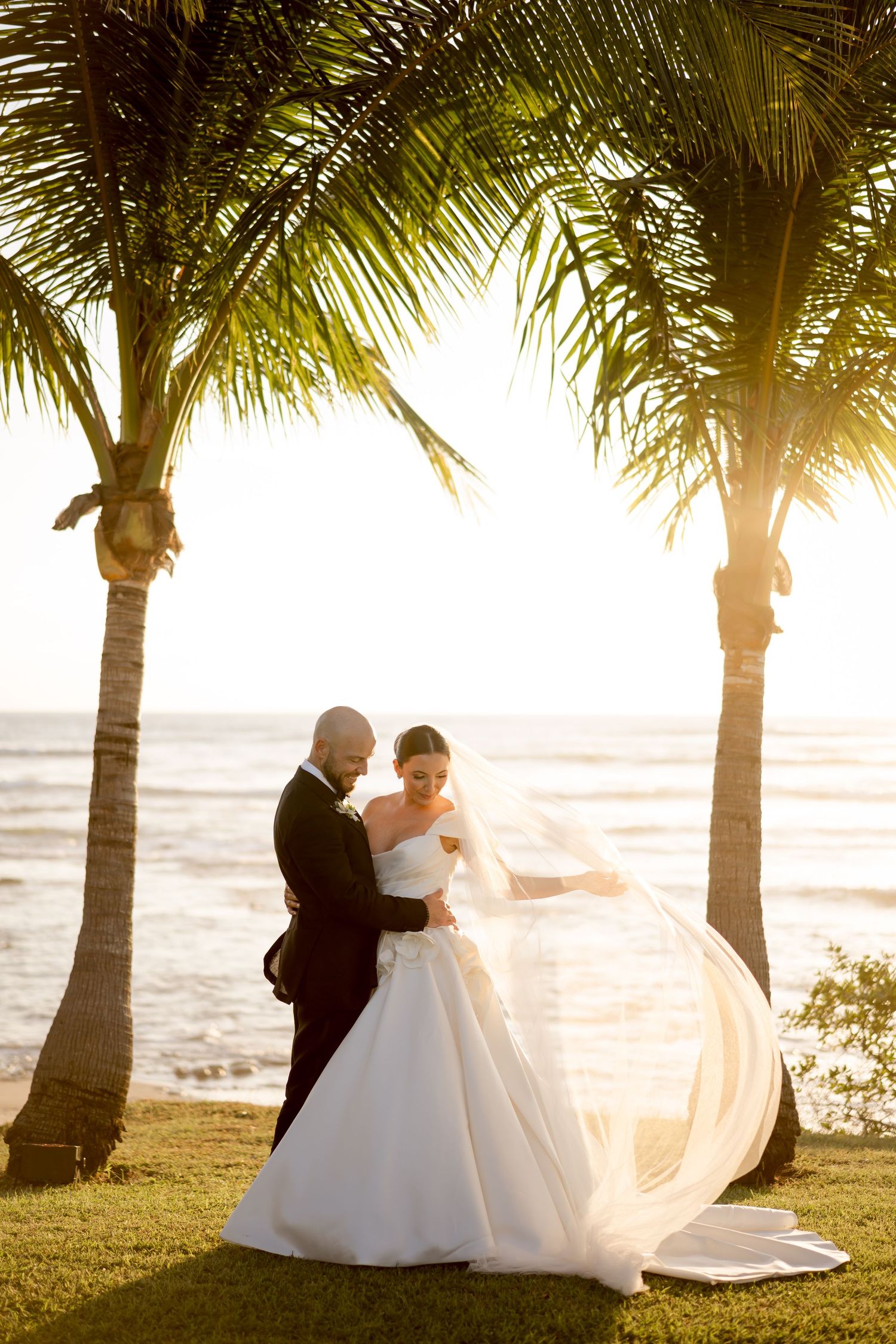 Bride and groom embracing beneath swaying palm trees as the setting sun creates a dramatic backlit wedding portrait in Hacienda Pinilla.