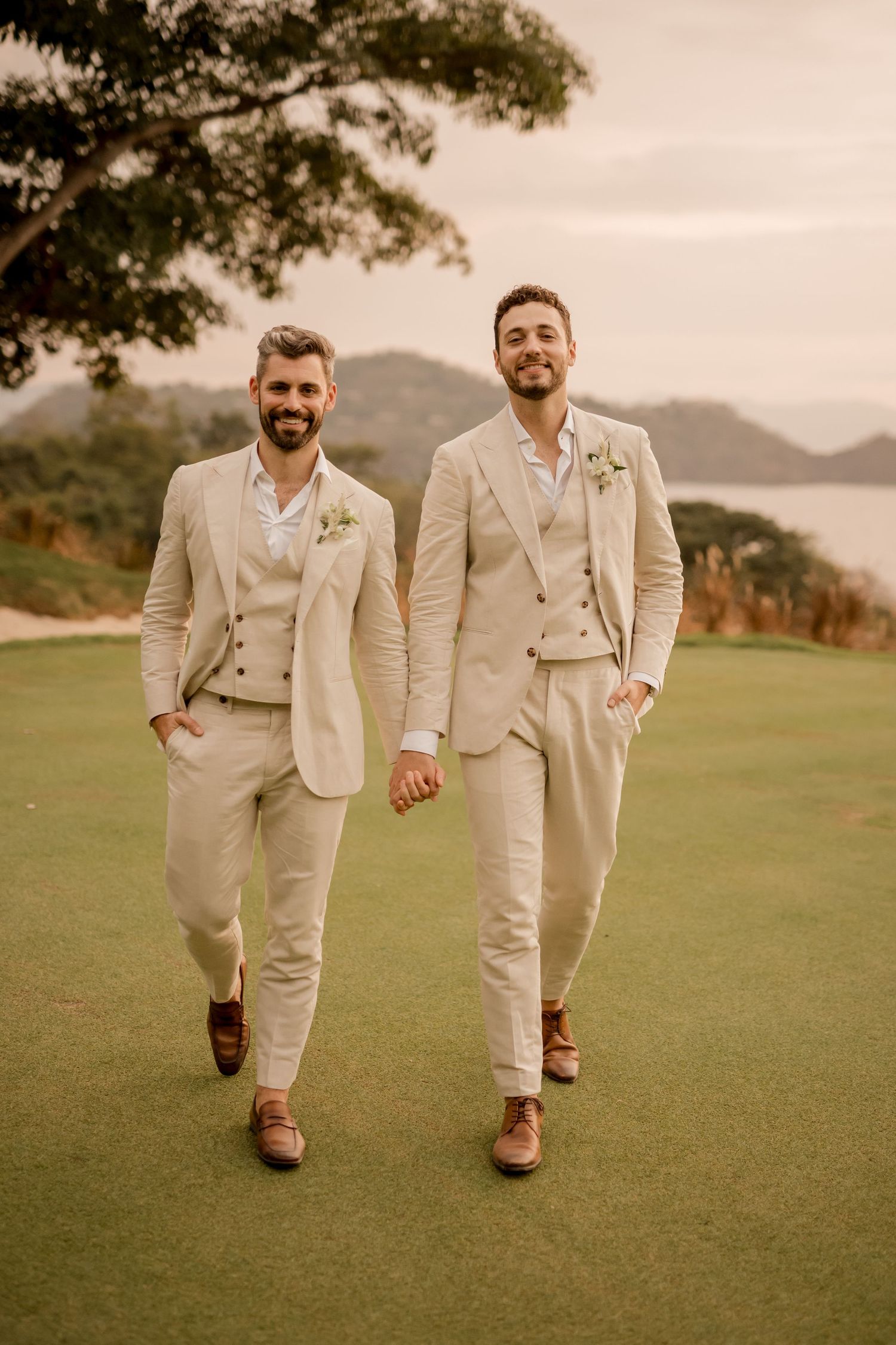 Two grooms in matching beige linen suits walk hand-in-hand on a golf course with ocean views on their wedding day in Papagayo, Costa Rica.