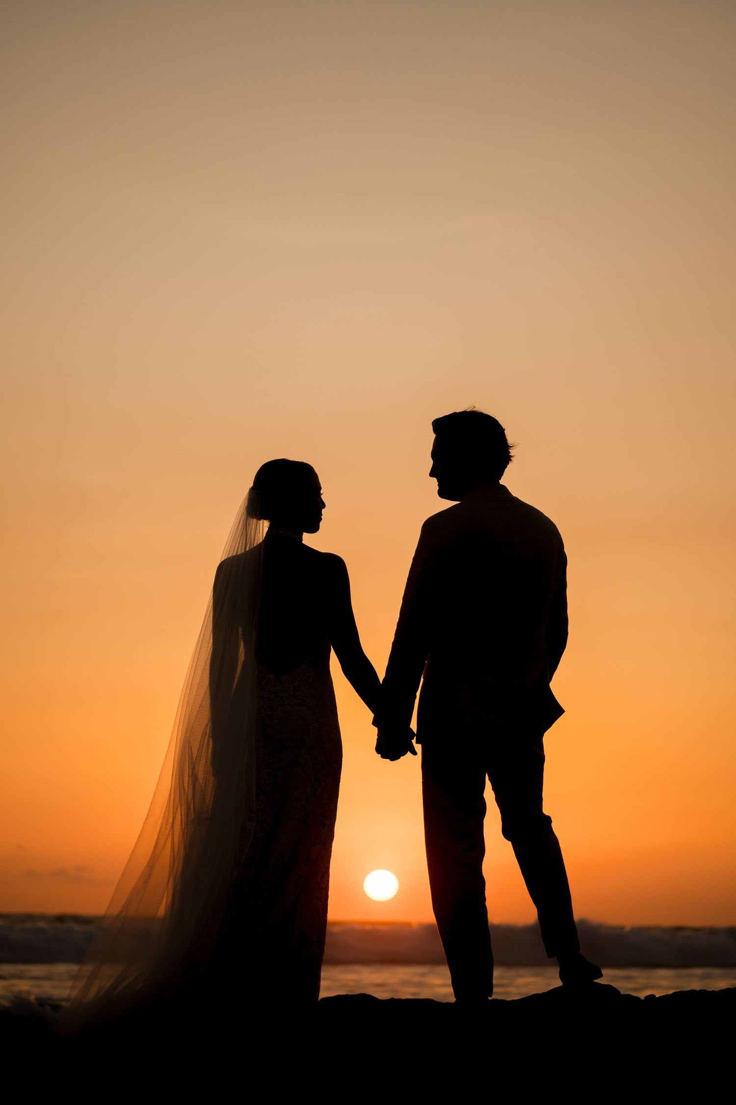 Silhouetted newlyweds holding hands as the sun sets over the Pacific Ocean during a destination wedding.
