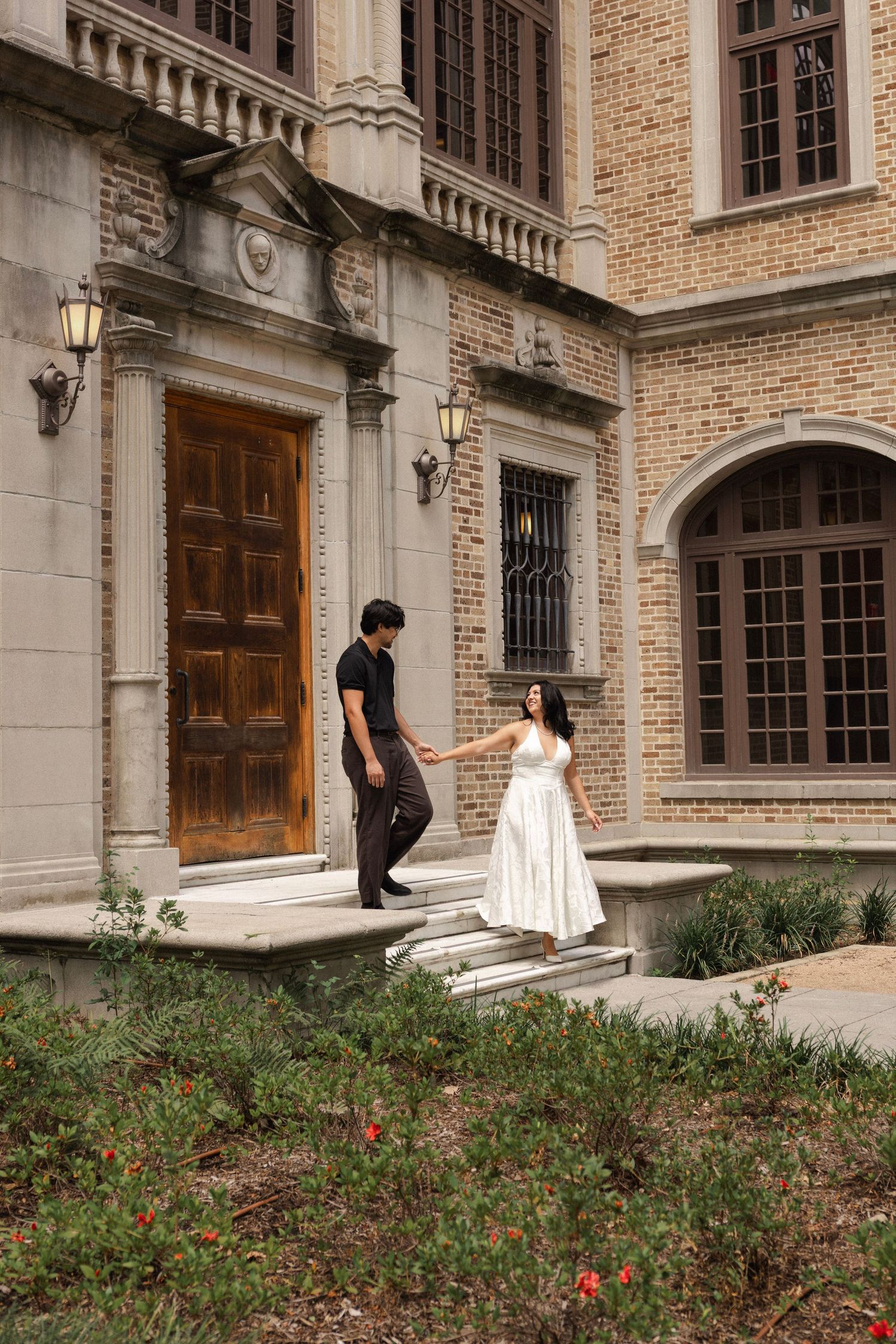 Couple holding hands on stone steps outside an elegant brick building with ornate wooden doors and arched windows.