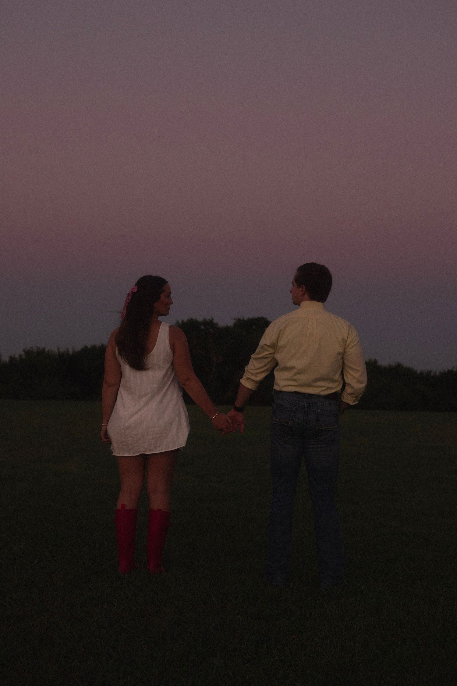 Couple holding hands in a field at dusk, woman in white dress and red boots, man in yellow shirt and jeans.