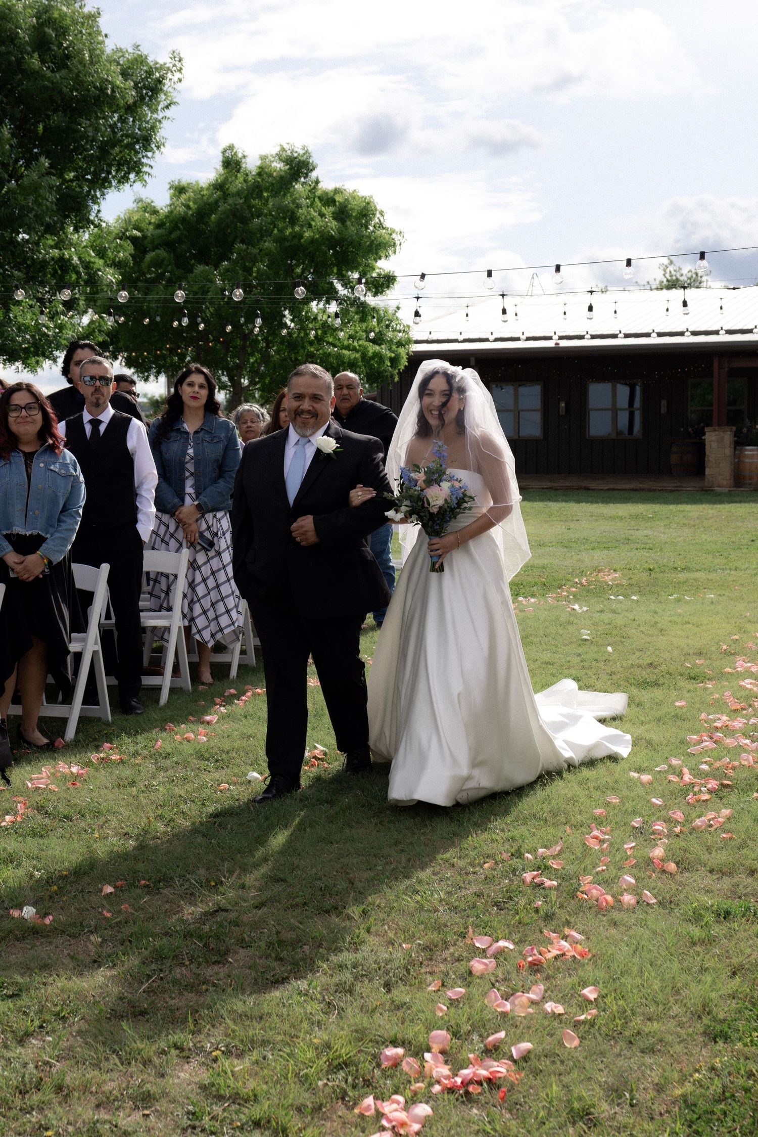 Father walks smiling bride down a rose petal-lined outdoor aisle as wedding guests look on.
