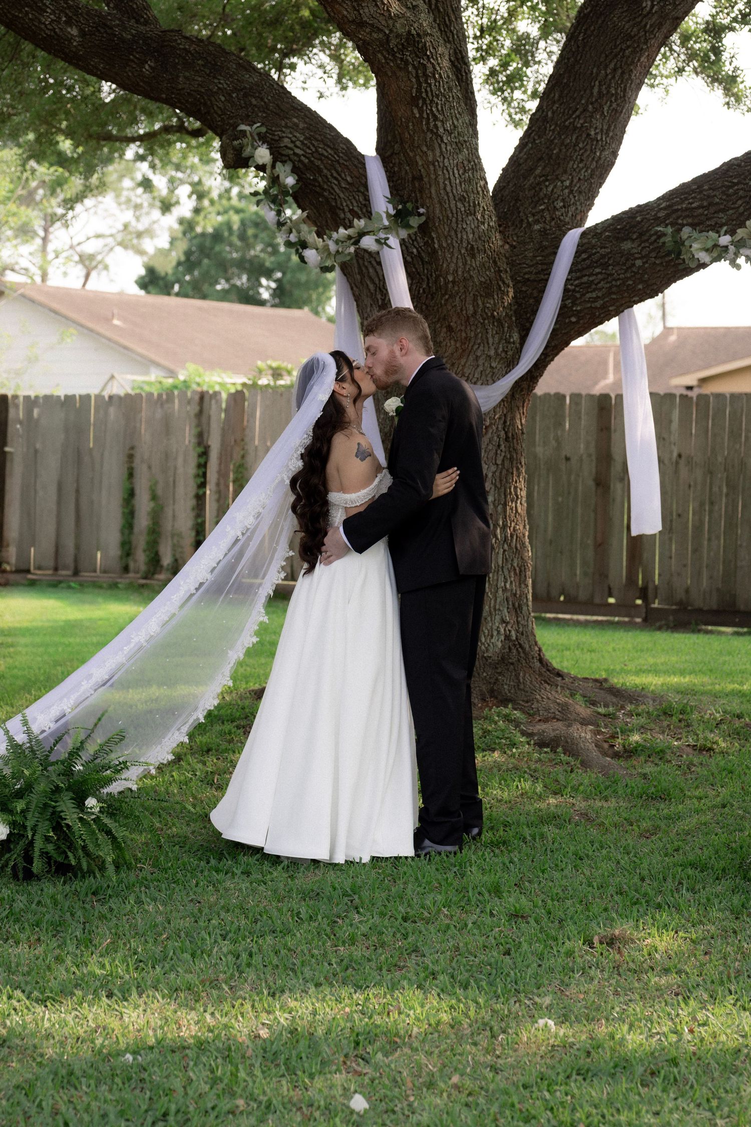 A bride and groom share their first kiss under a large tree at their backyard wedding.