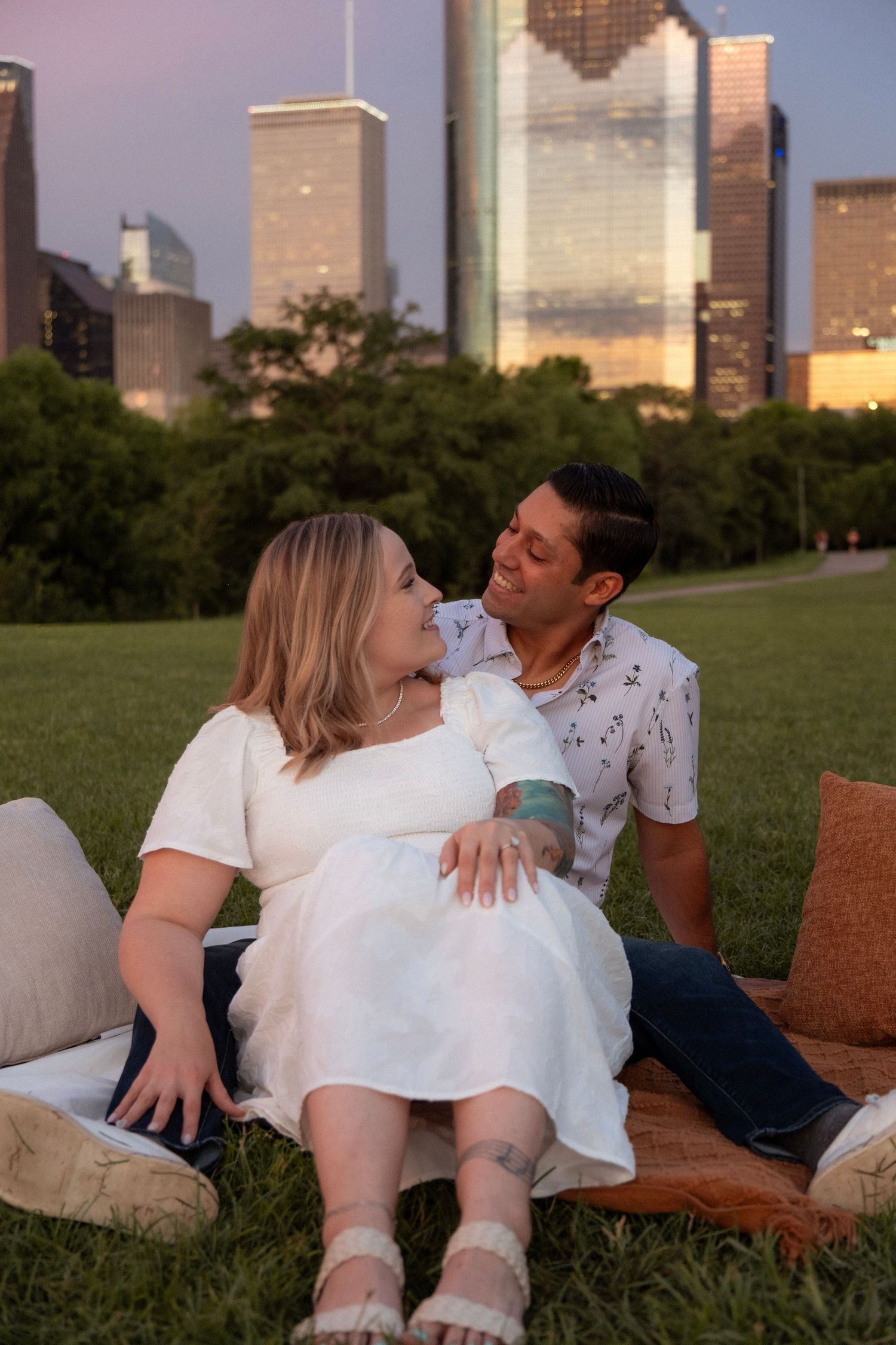 Pregnant couple sitting on grass blanket at sunset with Houston city skyline in background.