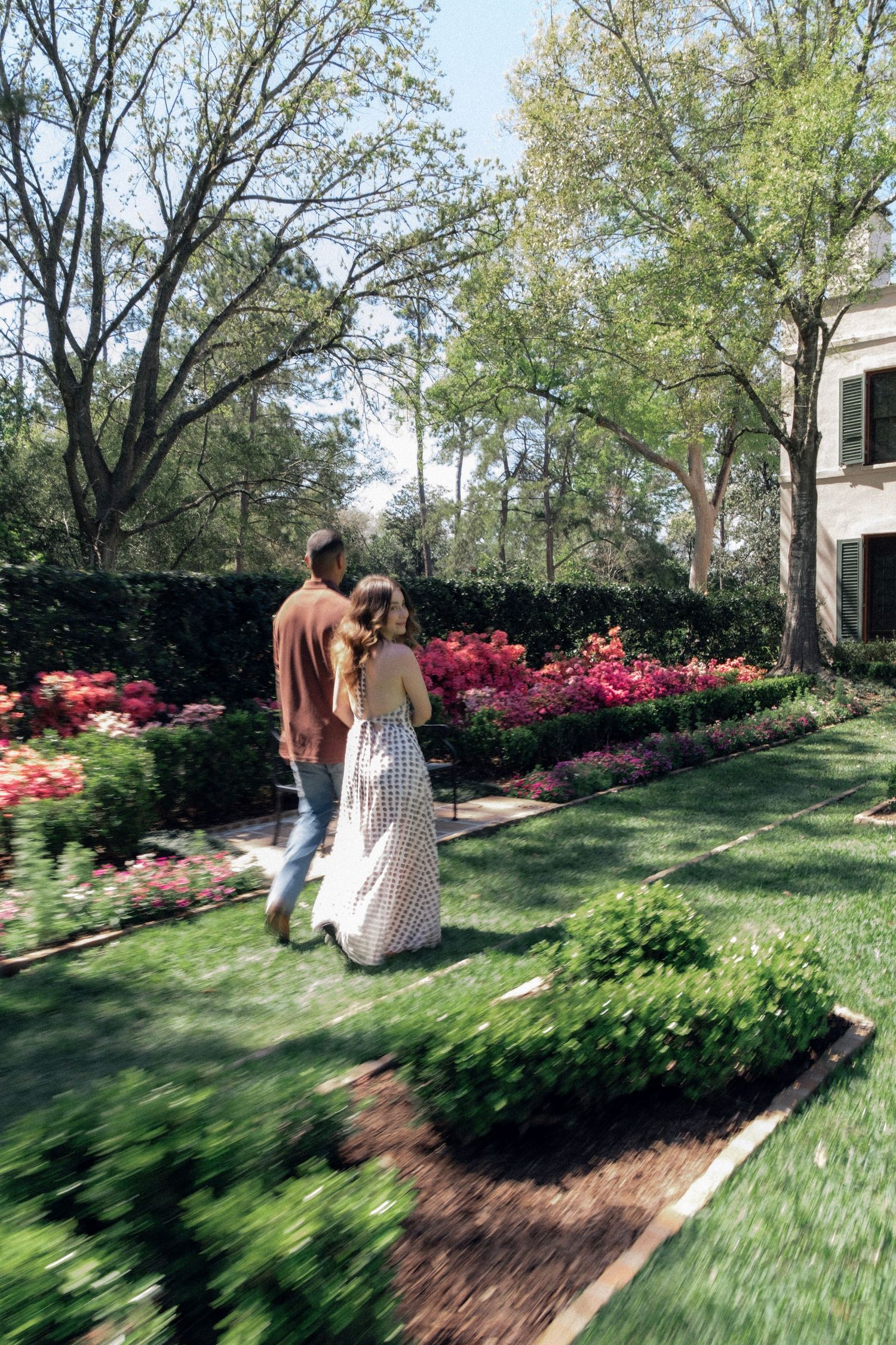 Couple walking together through lush garden with blooming pink and red flowers and green hedges.