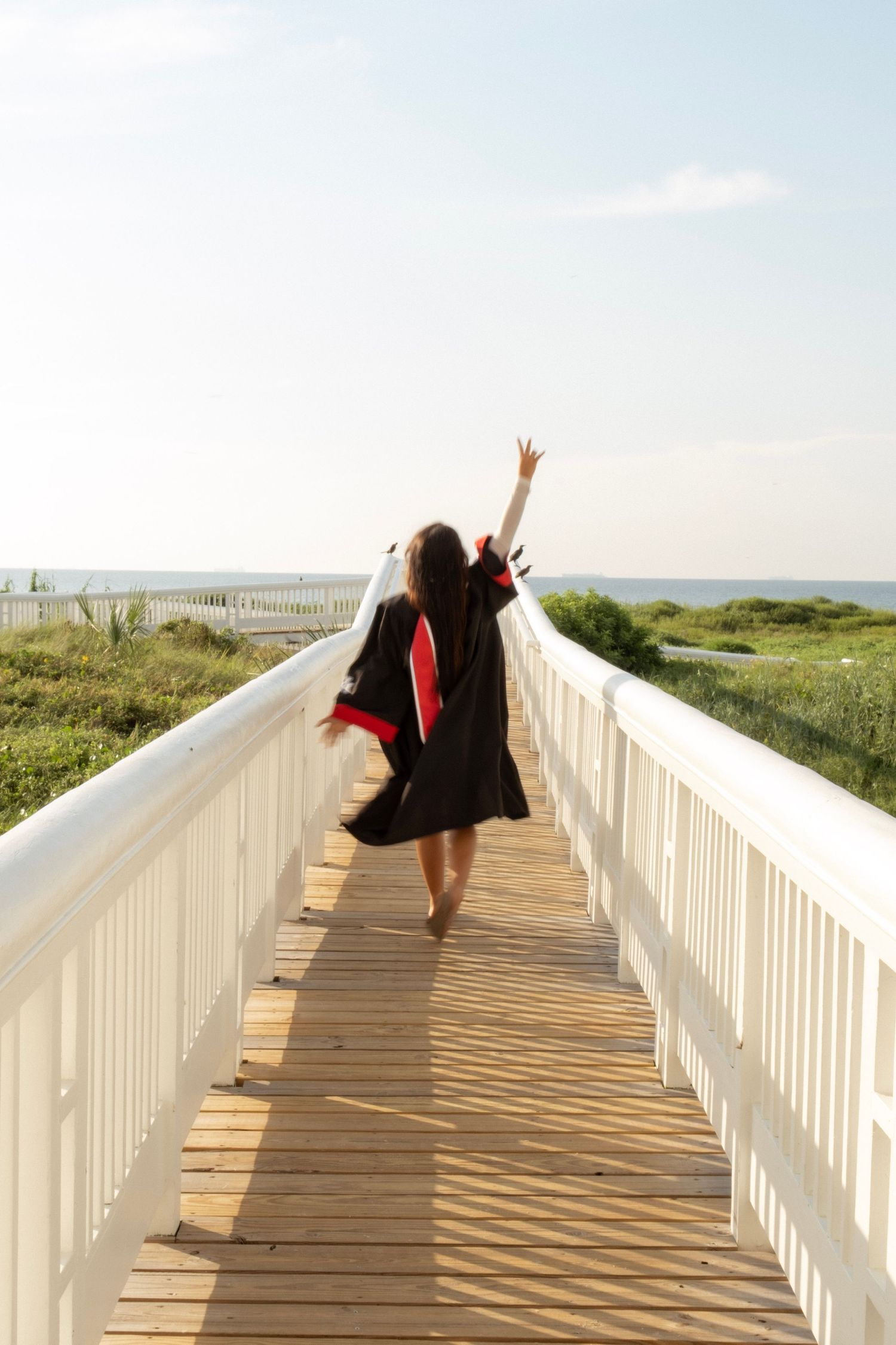 Graduate in black and red gown walking away on wooden boardwalk toward ocean with arm raised.