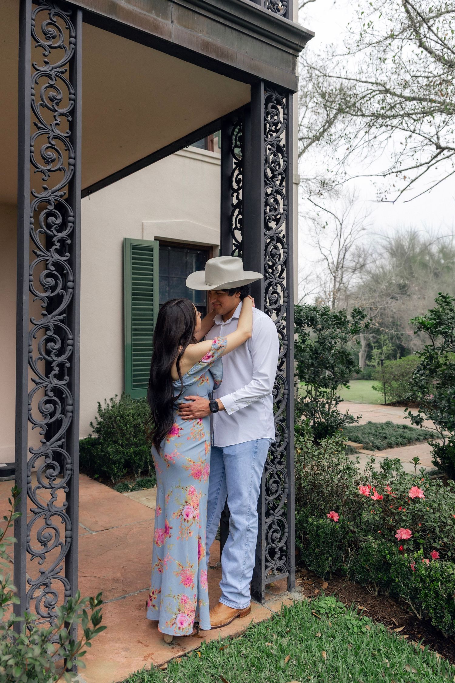 Couple embracing and kissing under ornate iron porch columns, woman in floral dress and man in cowboy hat.