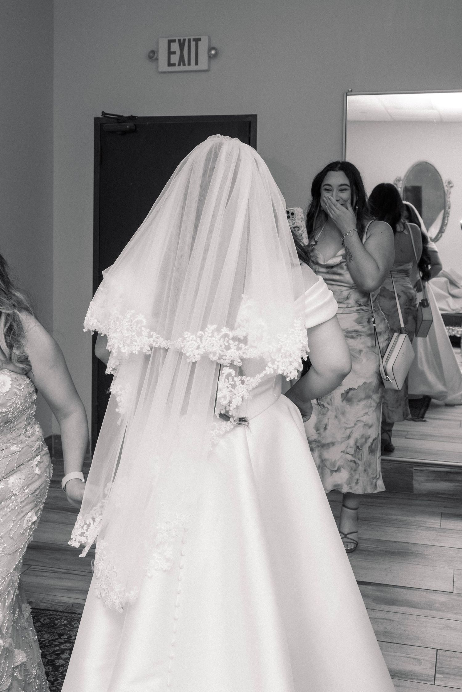 Black and white photo of a bride in a wedding gown and veil, with emotional guests reacting in a mirror-lined bridal suite.