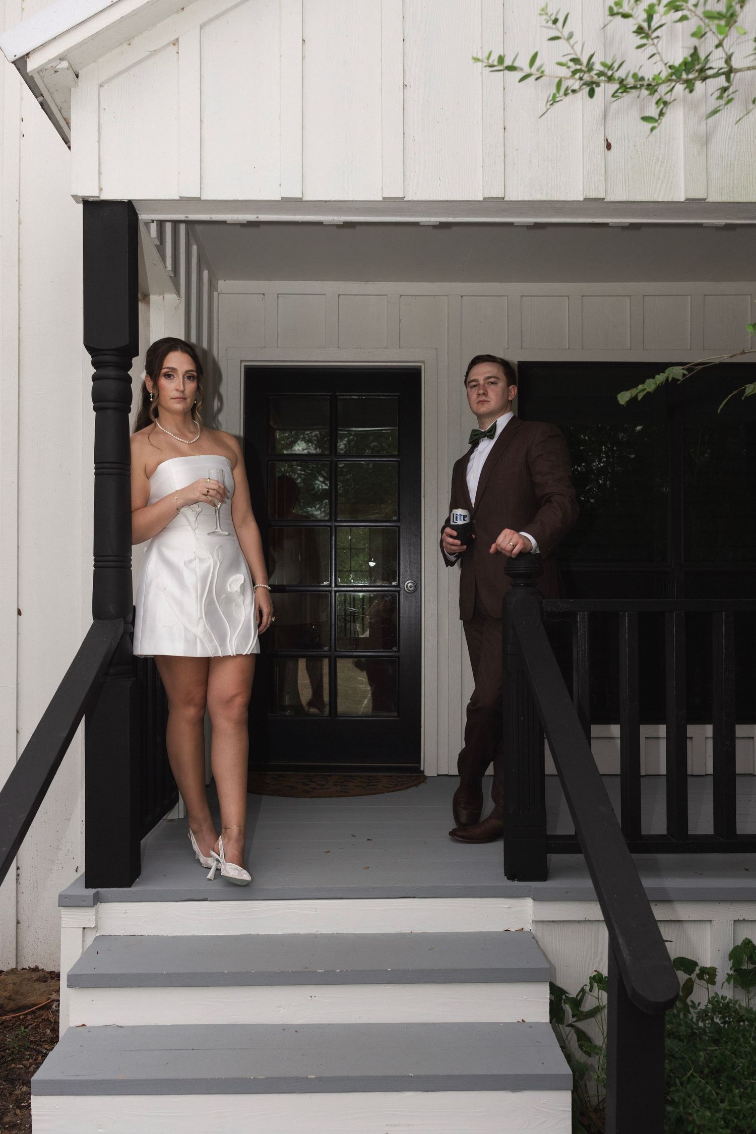 A couple in formal attire poses on porch steps, woman in white dress, man in brown suit with bow tie.