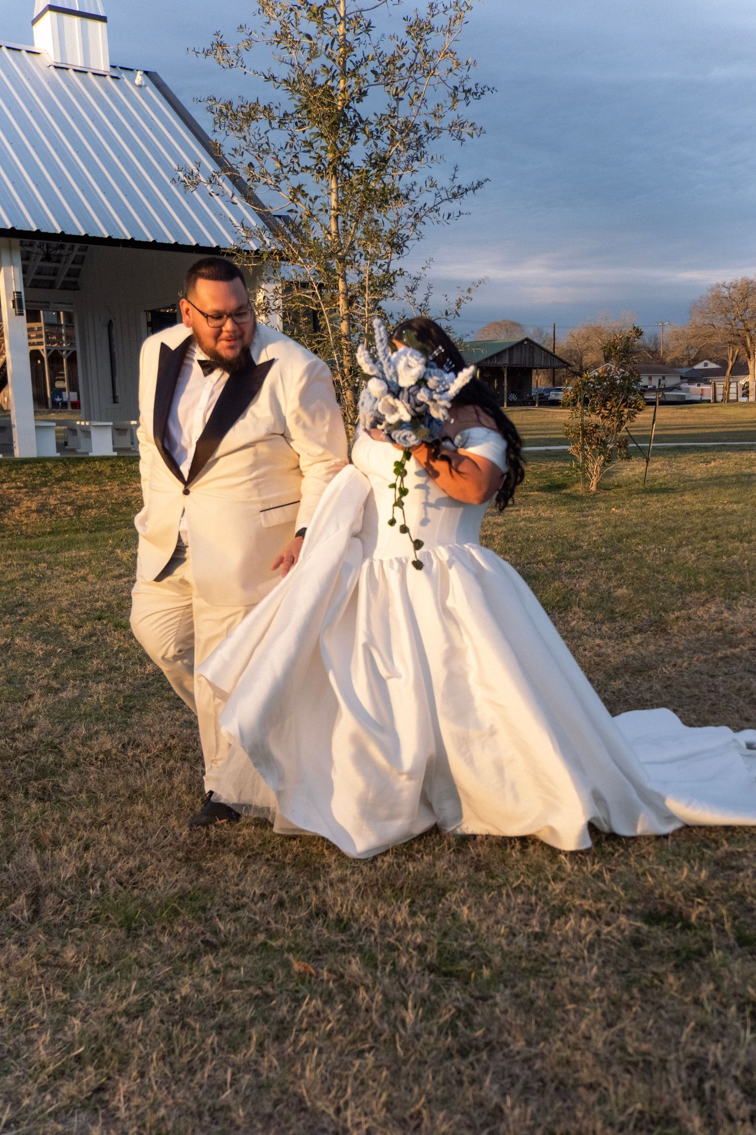Bride and groom pose outdoors in wedding attire, bride holding bouquet in white ball gown, groom in white tuxedo.