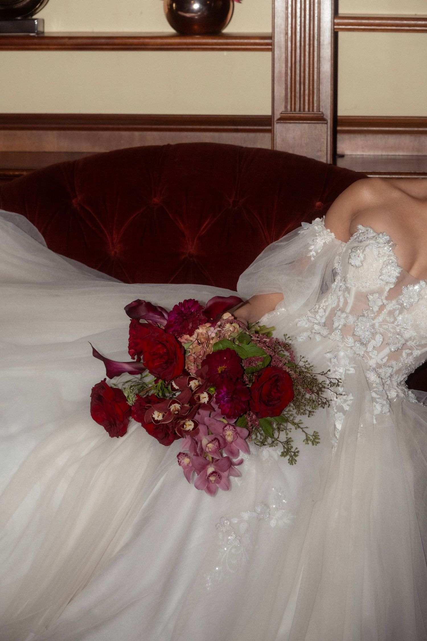 Bride in strapless lace ballgown holding stunning red and pink floral bouquet on ornate sofa.